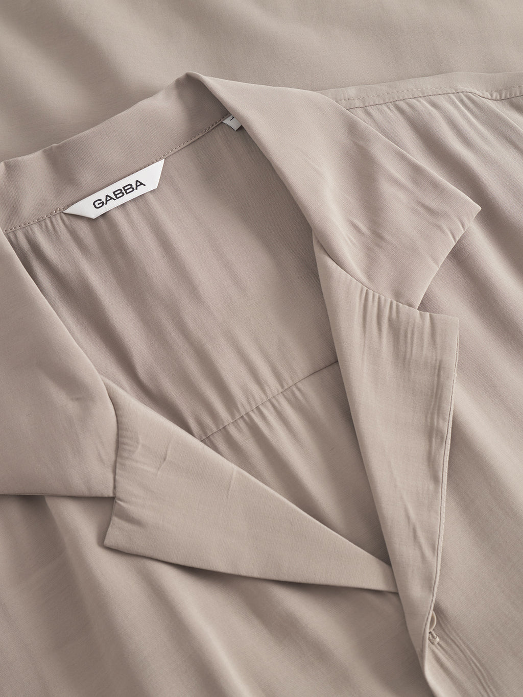 Close-up of the Jens Over Resort Yari SS in Sand Greige by Gabba, featuring a smooth light taupe fabric with a pointed collar. Delicate stitching details enhance the overall design, and a statement chest pocket adds flair.