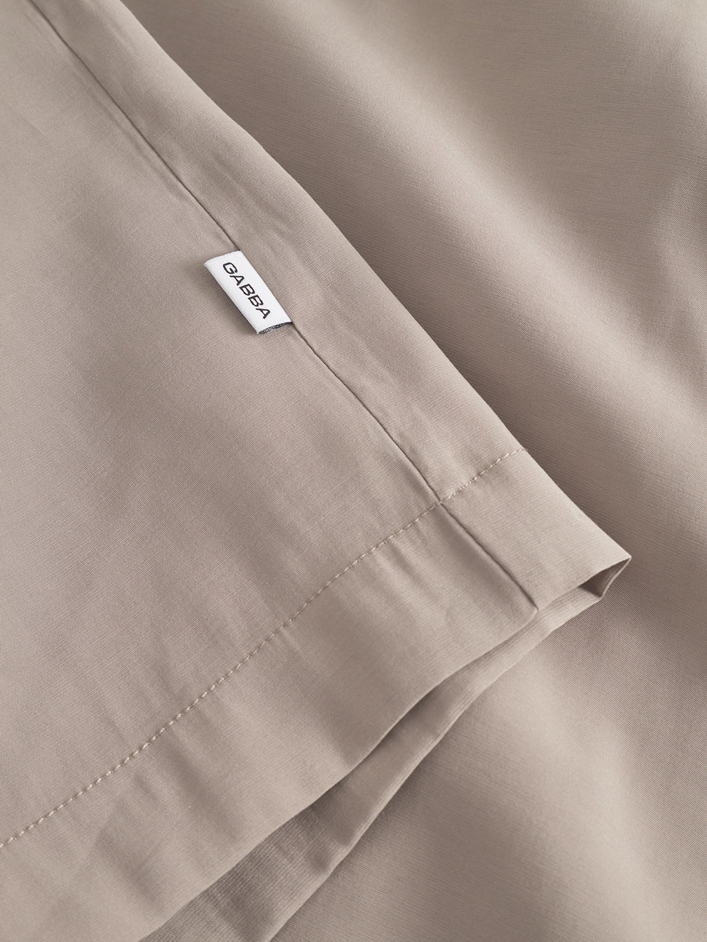 Close-up of Jen Over Resort Yari SS in Sand Greige by Gabba, featuring light beige sheets with a visible seam and a small white CALEEA tag. The premium fabric is soft with a smooth texture and minimalist design, evoking the elegance of an oversized shirt.