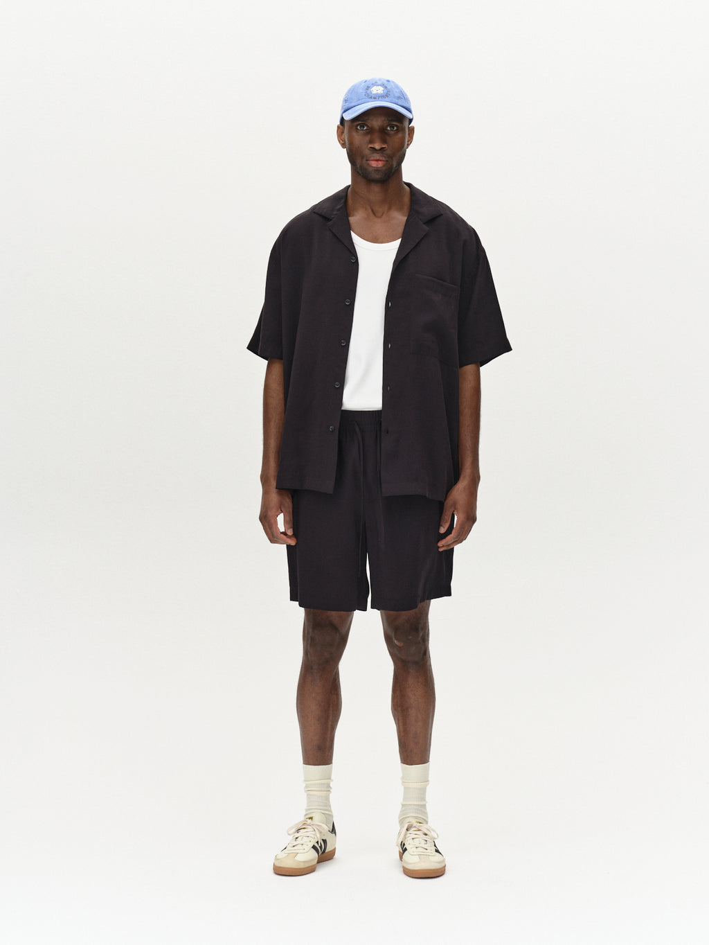 A man stands against a plain light background, wearing a light blue cap and a white tank top under a Gabba Jens Over Resort Yari SS - Meteorite oversized black open shirt with a statement chest pocket. He pairs it with black shorts, cream socks, and sneakers with dark accents.
