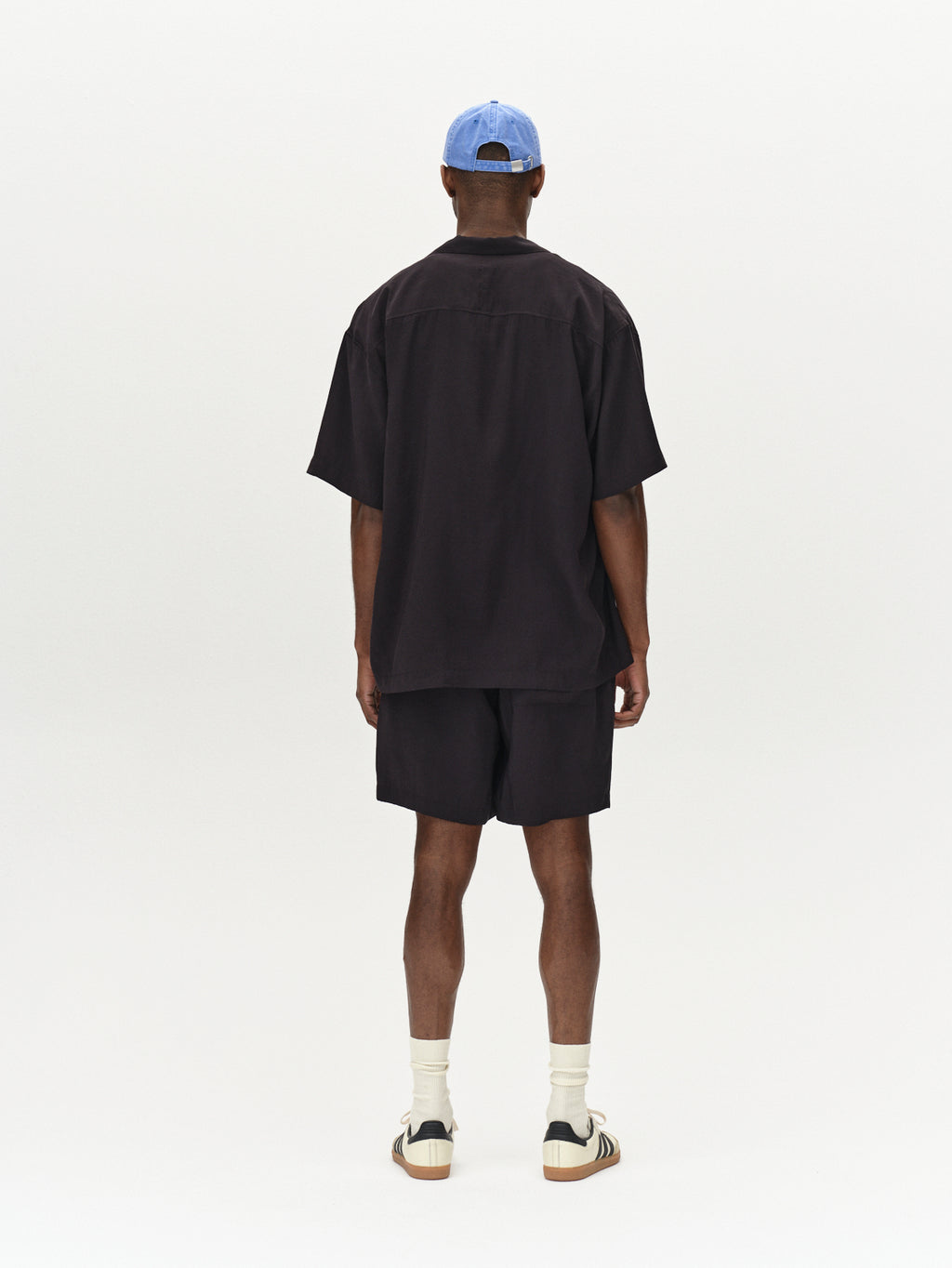 A person stands facing away, wearing the Jens Over Resort Yari SS - Meteorite by Gabba; its an oversized shirt with a statement chest pocket. The outfit includes matching shorts, white socks, and sneakers against a plain, light-colored background.
