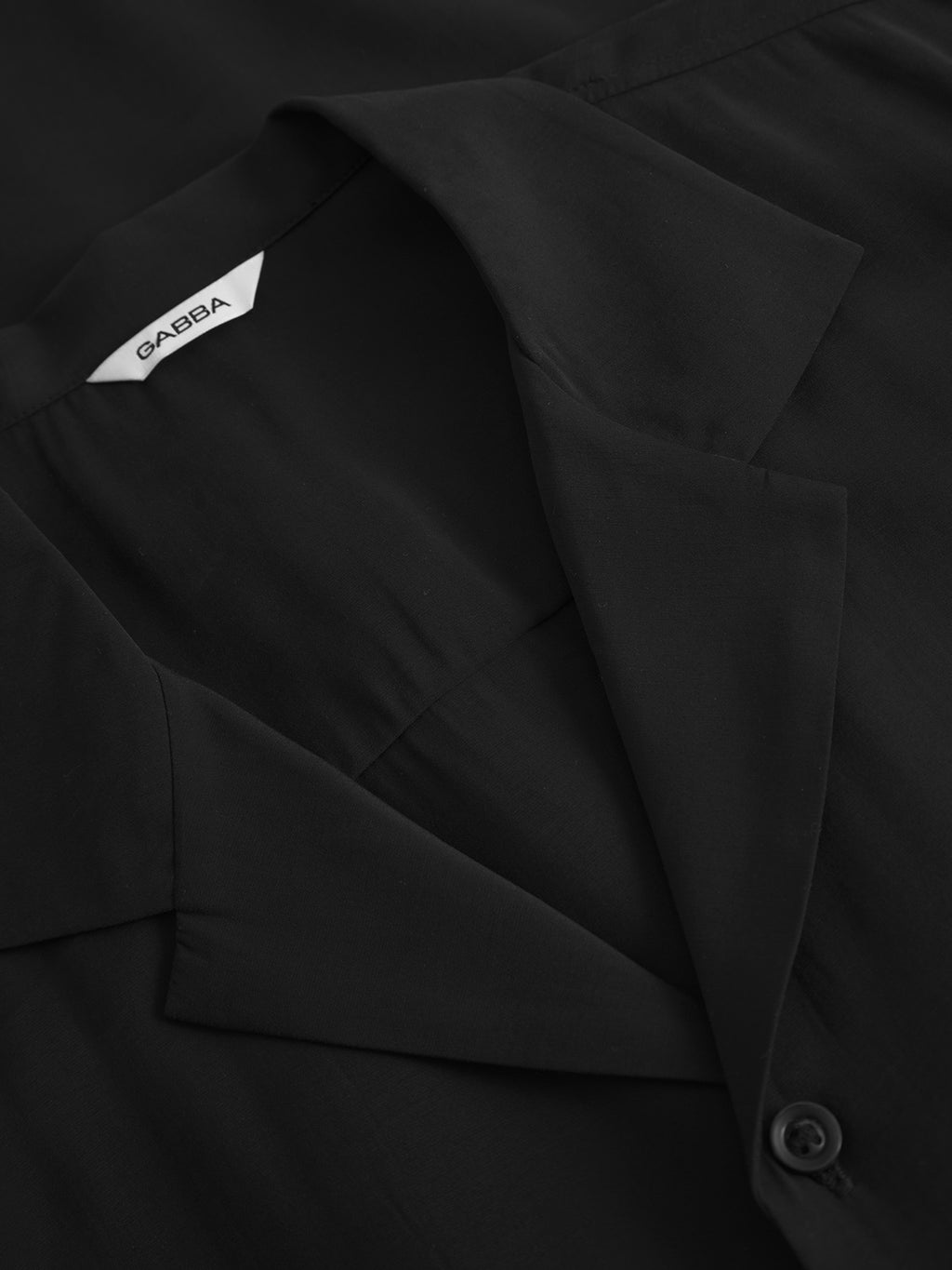 Close-up of Gabbas Jens Over Resort Yari SS - Meteorite. The black collared shirt features a brand tag and a partially visible button, with smooth, slightly shiny fabric exuding quality.