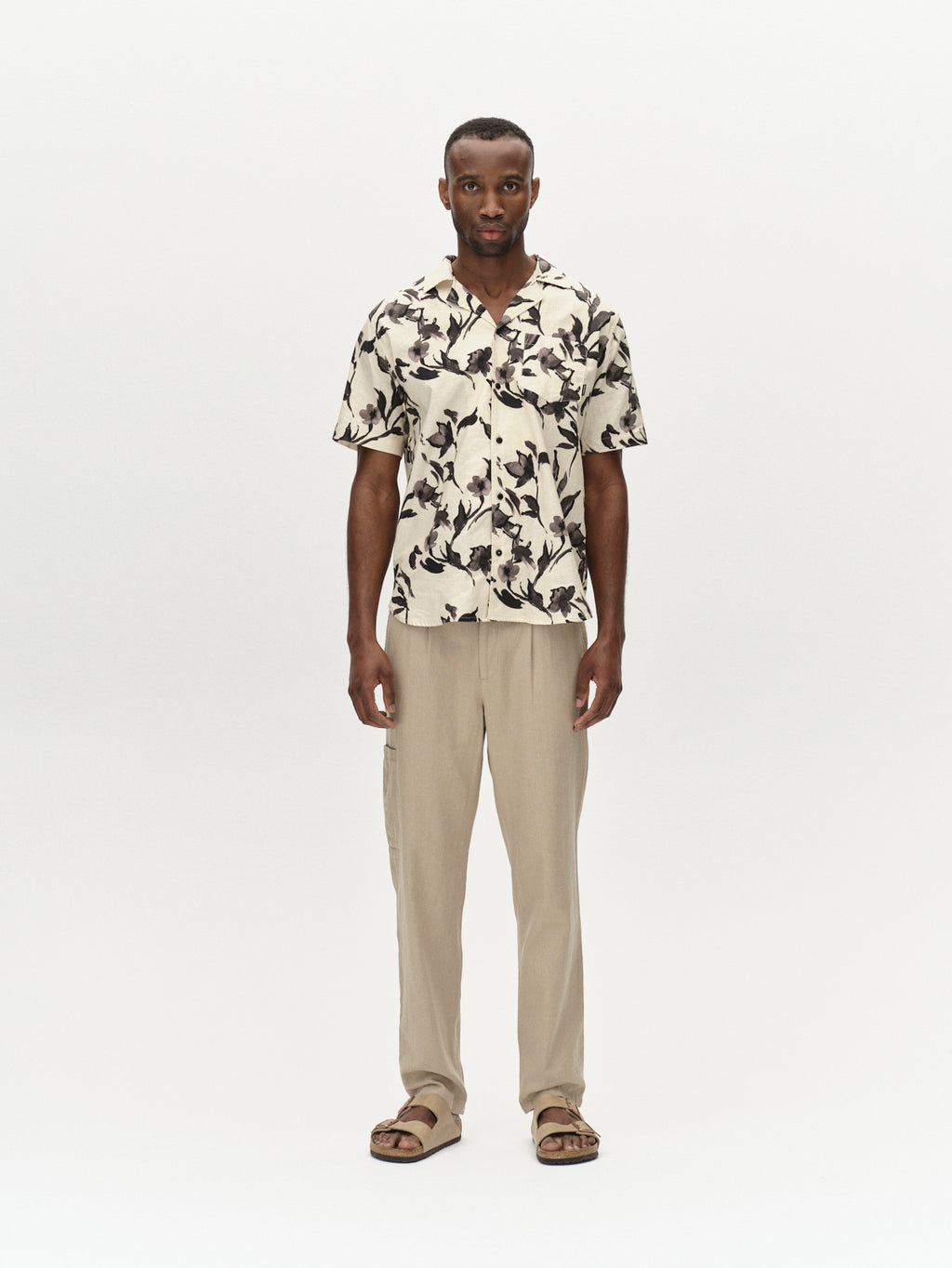 A man exudes a relaxed vibe against a plain background, wearing a short-sleeve shirt with a black and white abstract pattern, Gabbas beige Napoli 19 Side Lino - Sand Plaza pants, and brown sandals. A watch adorns his left wrist as he maintains a neutral expression.