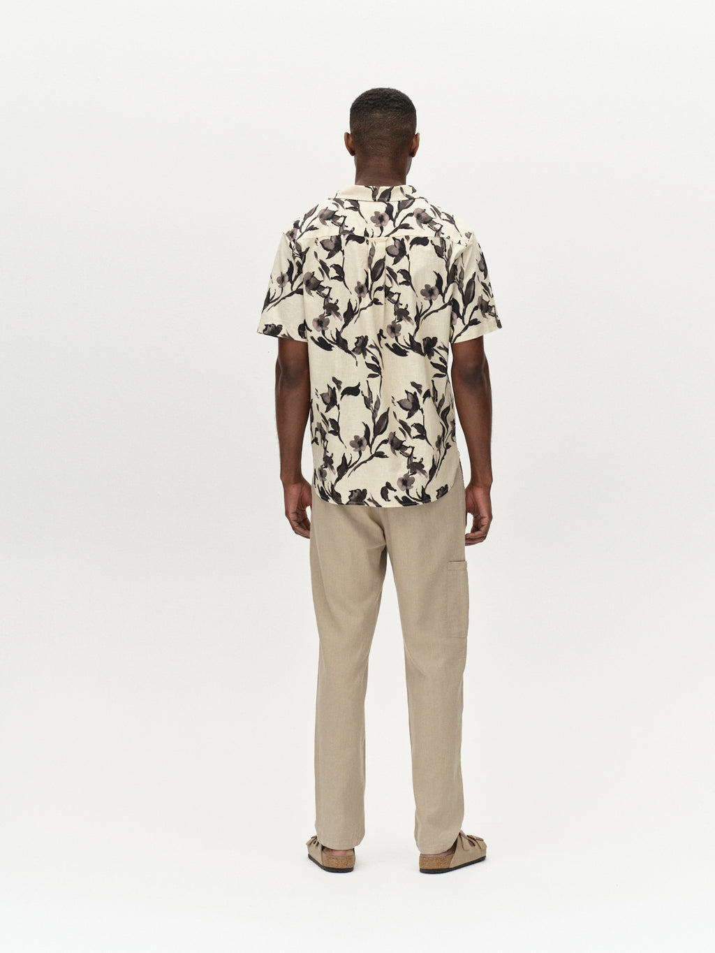 Against a plain backdrop, a person exudes relaxation in a short-sleeved shirt featuring black leaf patterns on cream and pairs it with Gabbas Napoli 19 Side Lino Sand Plaza pants and sandals, highlighting an effortless blend of style and comfort.