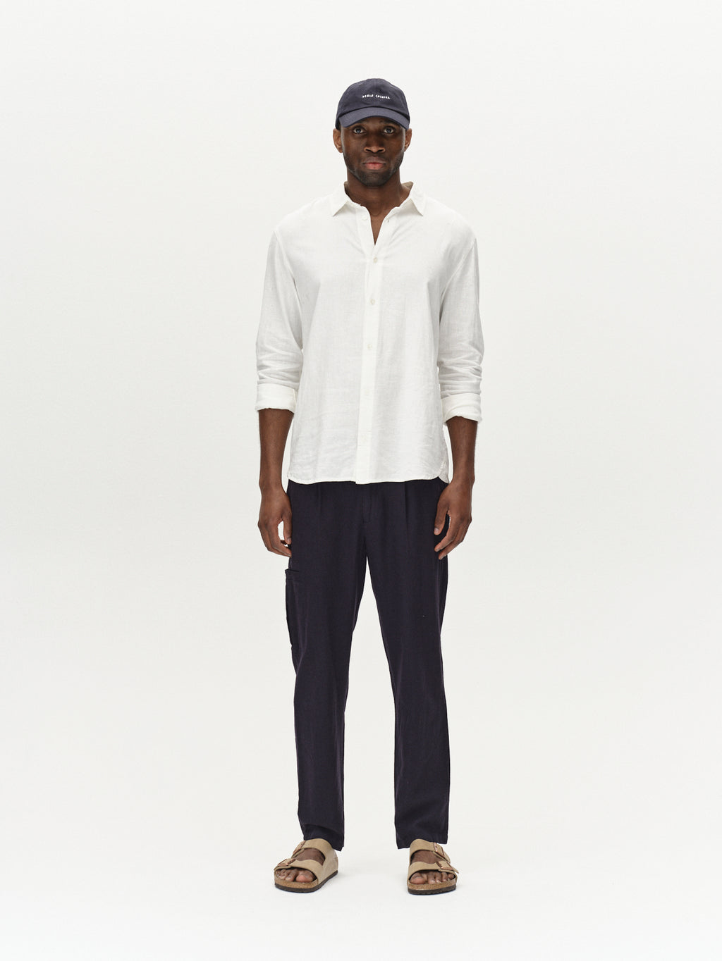 A person exudes a relaxed vibe wearing a white button-up shirt, Gabbas Napoli 19 Side Lino pants in Night Sky, with beige sandals and a matching cap, standing casually against a plain white background.