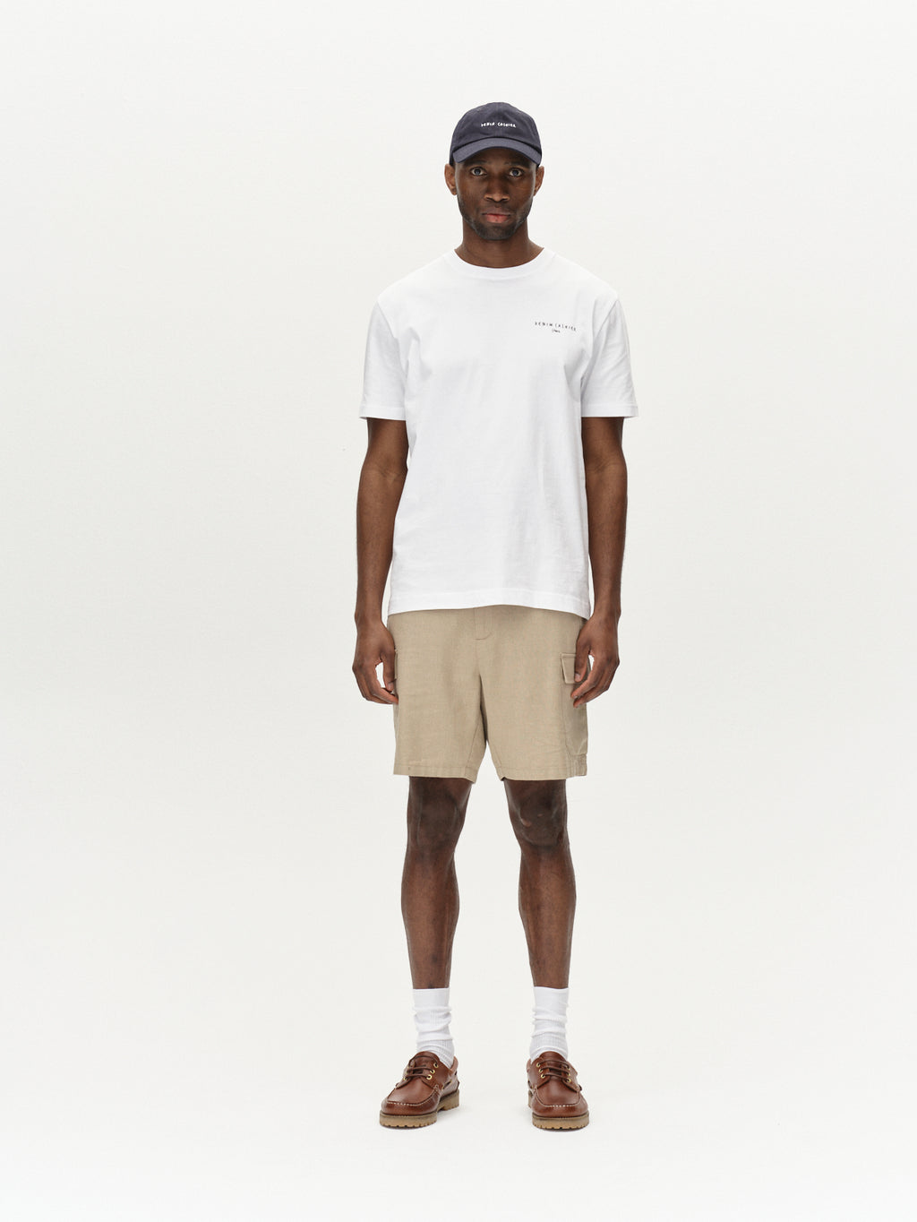 Against a plain backdrop, a person wears a navy cap and white t-shirt paired with Gabbas Alfa Cargo Lino - Sand Plaza beige linen shorts. The look is completed with white socks and brown loafers, offering a lightweight, casual, and relaxed style.