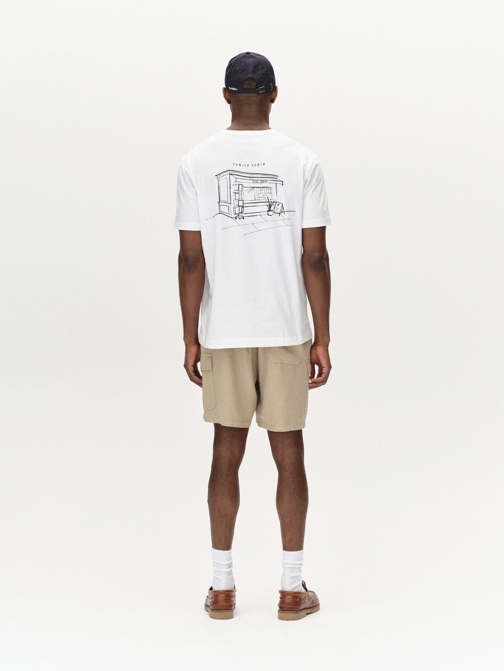 Rear view: A person wears a white sketch-design T-shirt, Alfa Cargo Lino - Sand Plaza shorts by Gabba for a breathable feel, white socks, brown sandals, and a dark cap against a plain white background.