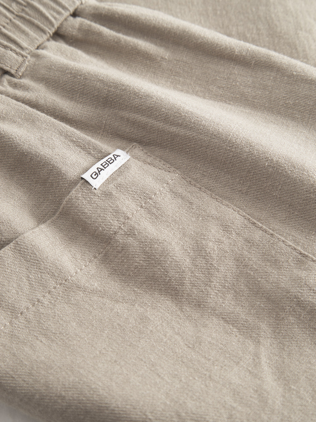 Close-up of beige Alfa Cargo Lino pants in Sand Plaza by Gabba, featuring an elastic waistband made from lightweight linen. A small white label with Gabba is stitched near the seam, highlighting the breathable fabrics comfort and casual style.