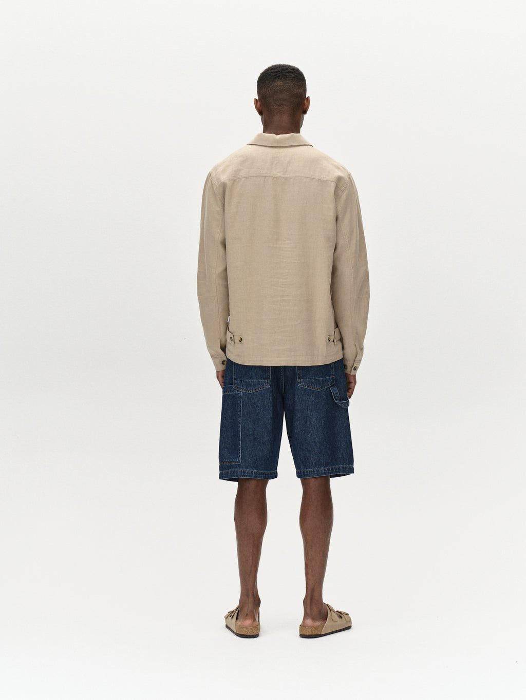 Against a plain white background, a person with short hair stands facing away, wearing Gabbas Peter Lino - Sand Plaza beige linen/cotton overshirt, denim shorts, and sandals.