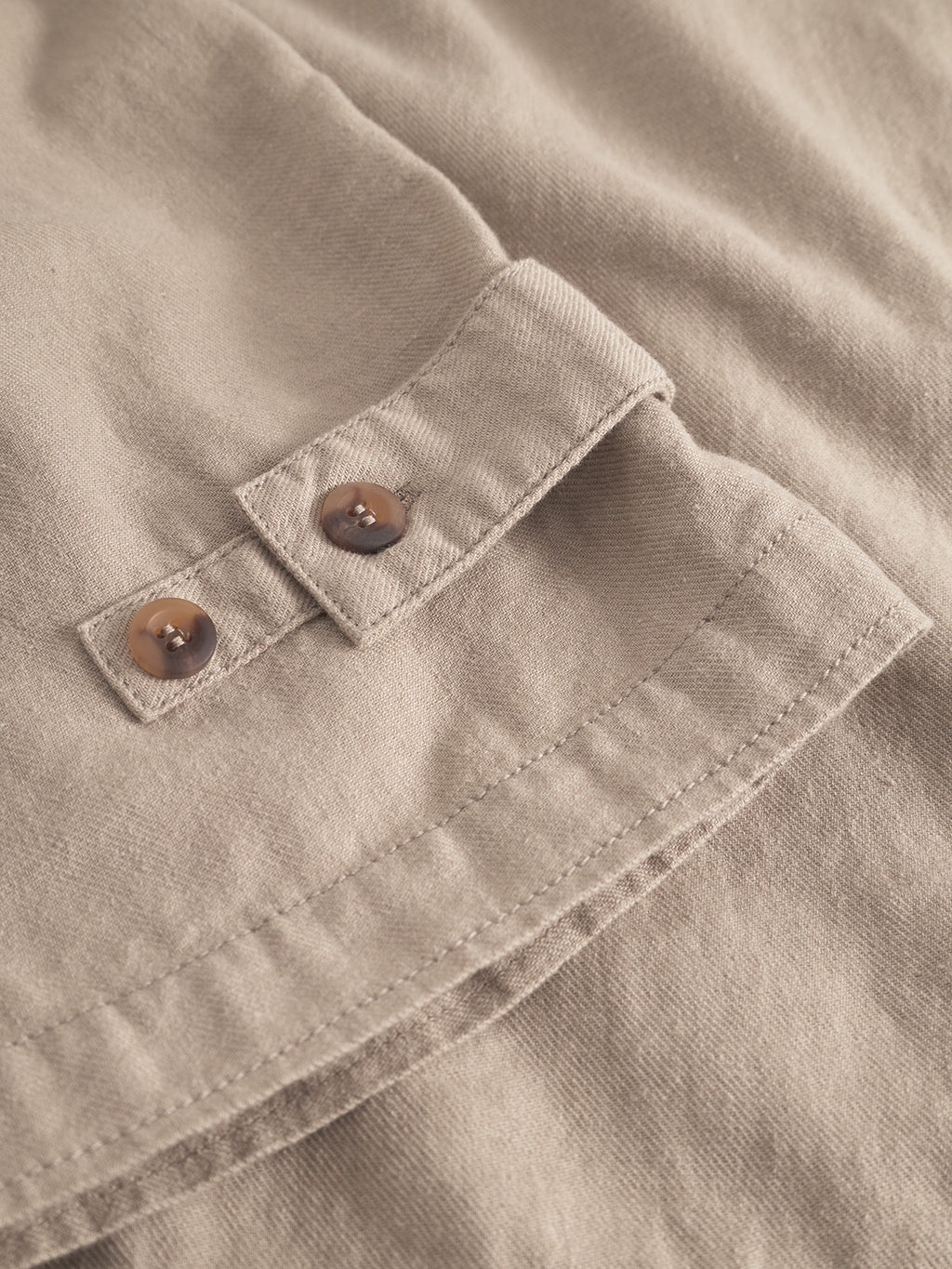 A close-up of Gabbas Peter Lino - Sand Plaza reveals a beige linen/cotton fabric with two brown buttons on a strap, showcasing neat stitching and a soft, matte finish. This design suggests an overshirt or accessory perfect for spring.