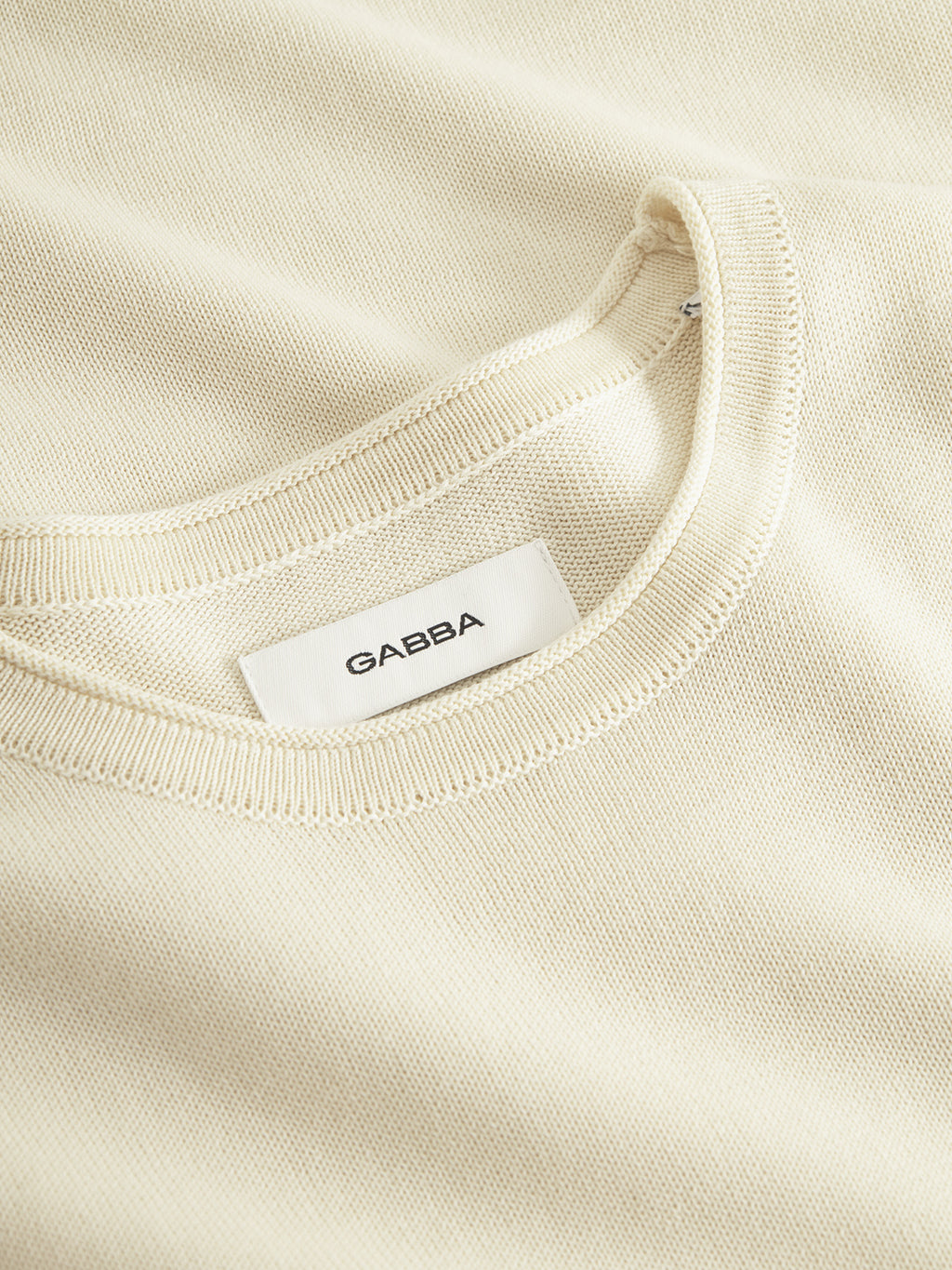Close-up of the Bela Fix LS - Ecru Light by Gabba, showcasing a cream-colored premium knit fabric with a visible neckline and attached label. Made from 100% cotton, it offers superior comfort with its soft texture and embodies a simple minimalist design.
