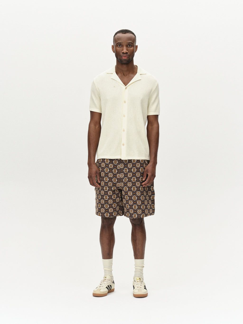 Against a white background, someone wears Gabbas Geno Resort SS in Ecru Light with patterned shorts and sneakers. The retro vibe is enhanced by the fabrics subtle texture, while their arms rest casually at their sides, capturing effortless style.