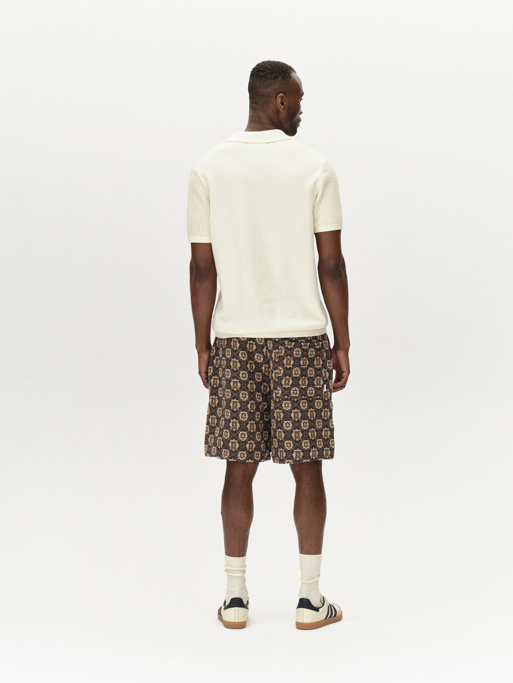 Facing away, a person embraces a retro vibe in the Geno Resort SS - Ecru Light by Gabba, styled with patterned shorts. The ensemble is completed with white socks and beige sandals, set against a plain white background.