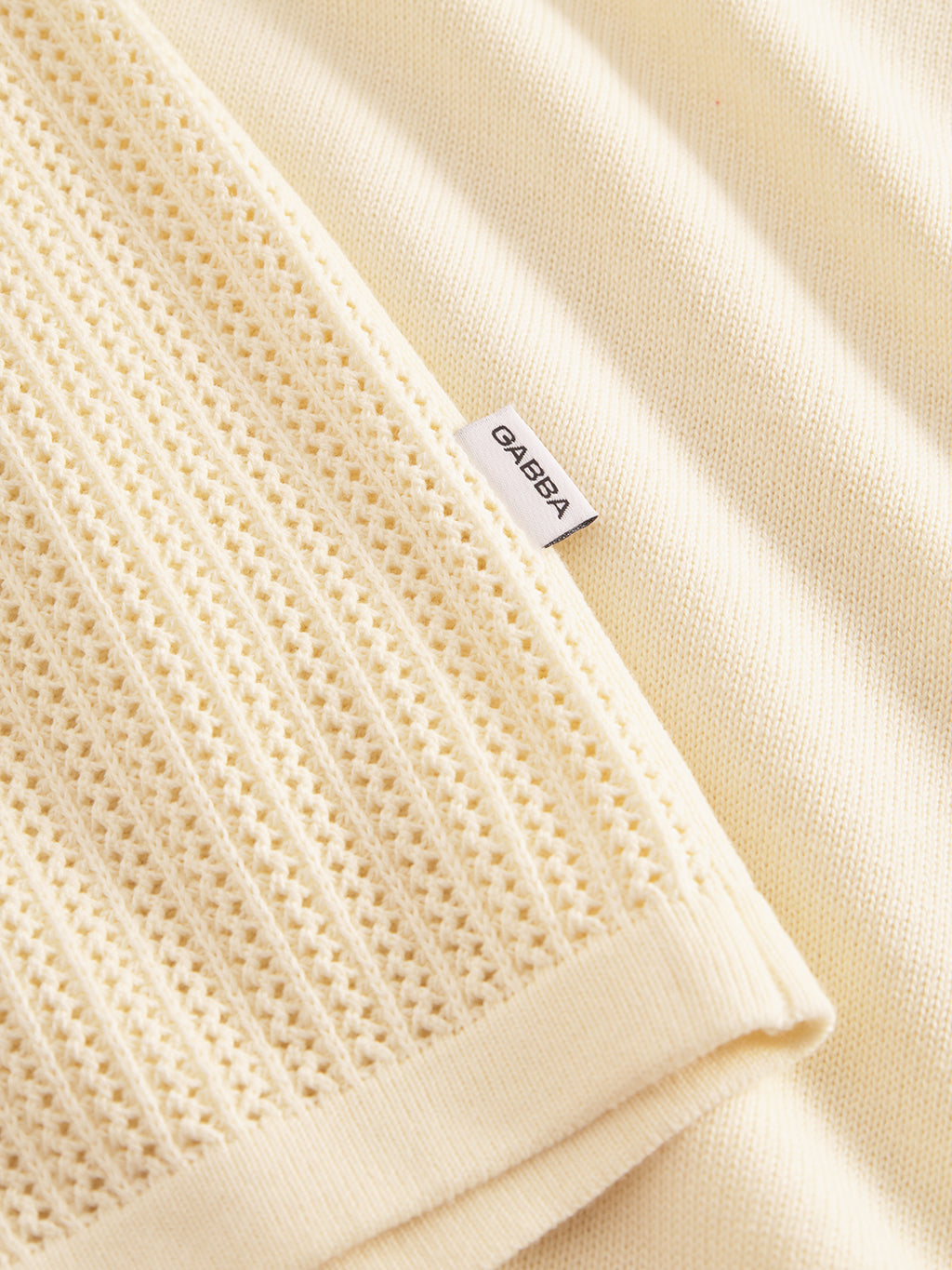 Close-up of Geno Resort SS - Ecru Light by Gabba: Cream fabric with smooth and premium knit diamond textures. A white tag reading Gabba is near the textured area. Soft, warm lighting highlights its retro vibe.