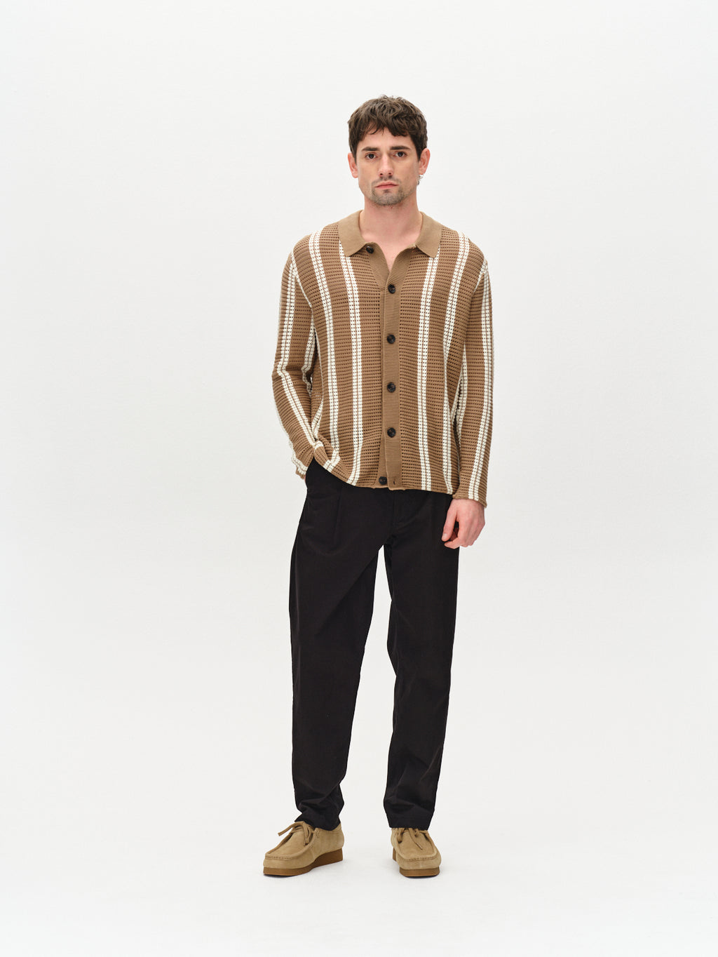 A person is on a plain background, wearing the Dezz Stripes LS by Gabba - a brown striped long-sleeve shirt, black pants, and tan shoes. They have short brown hair, a neutral expression, and hands in pockets.