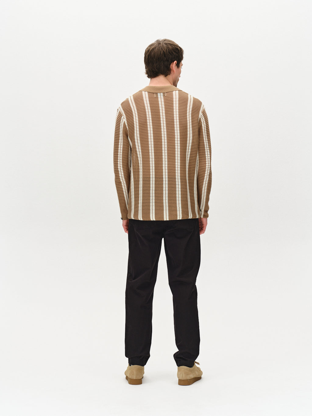 A person faces away in the Dezz Stripes LS by Gabba, showcasing its brown and white vertical stripes. They wear black pants and beige shoes against a plain white background.
