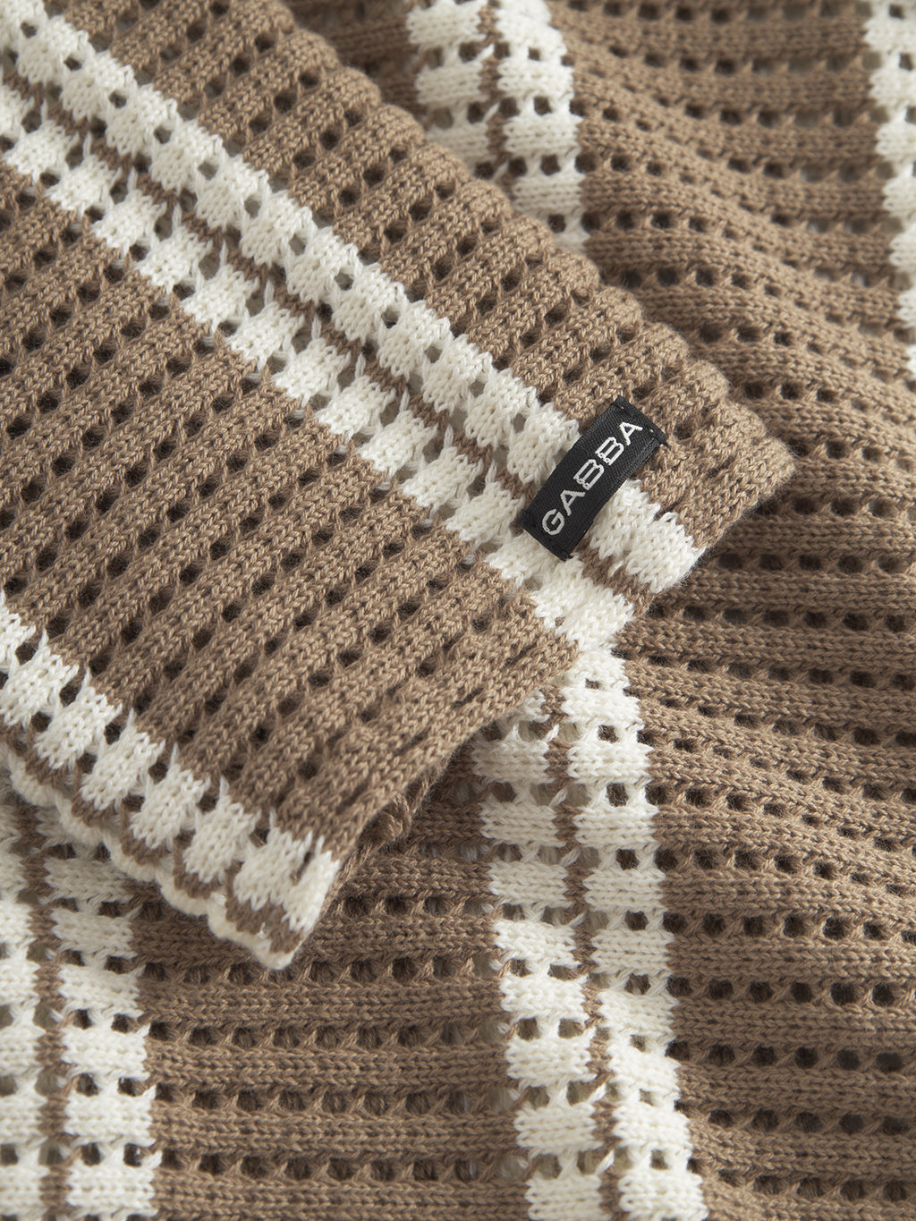 Close-up of textured Dezz Stripes LS fabric by Gabba with brown and white vertical stripes, featuring a black label. The cotton material has a waffle-like pattern, ideal for cozy sweaters or blankets.