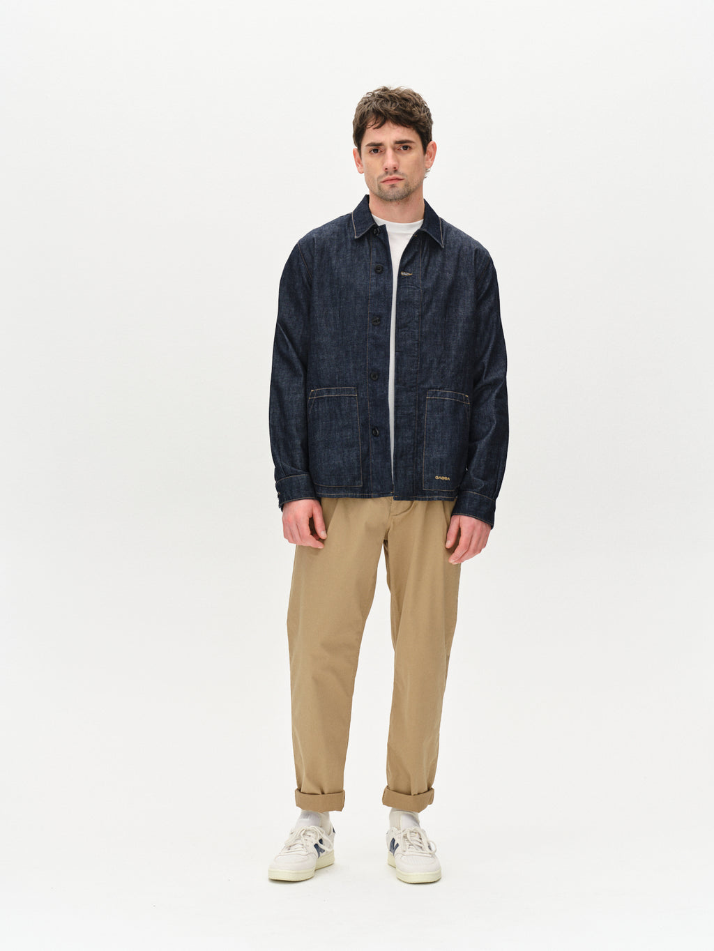 A man stands against a plain white backdrop wearing a Gabba Jap Beam dark blue denim jacket, a crisp white shirt, beige pants, and sleek white sneakers. He looks directly at the camera with a neutral expression.