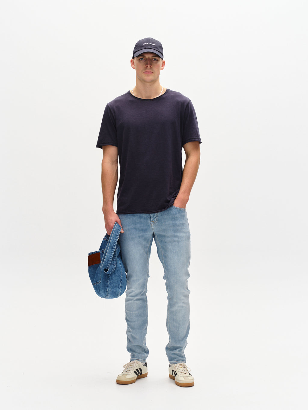 A person stands against a plain backdrop, wearing a black cap, dark t-shirt, slim silhouette jeans, and beige sneakers, holding a Gabba Jones Just - Lt. Blue Denim jacket in one hand.