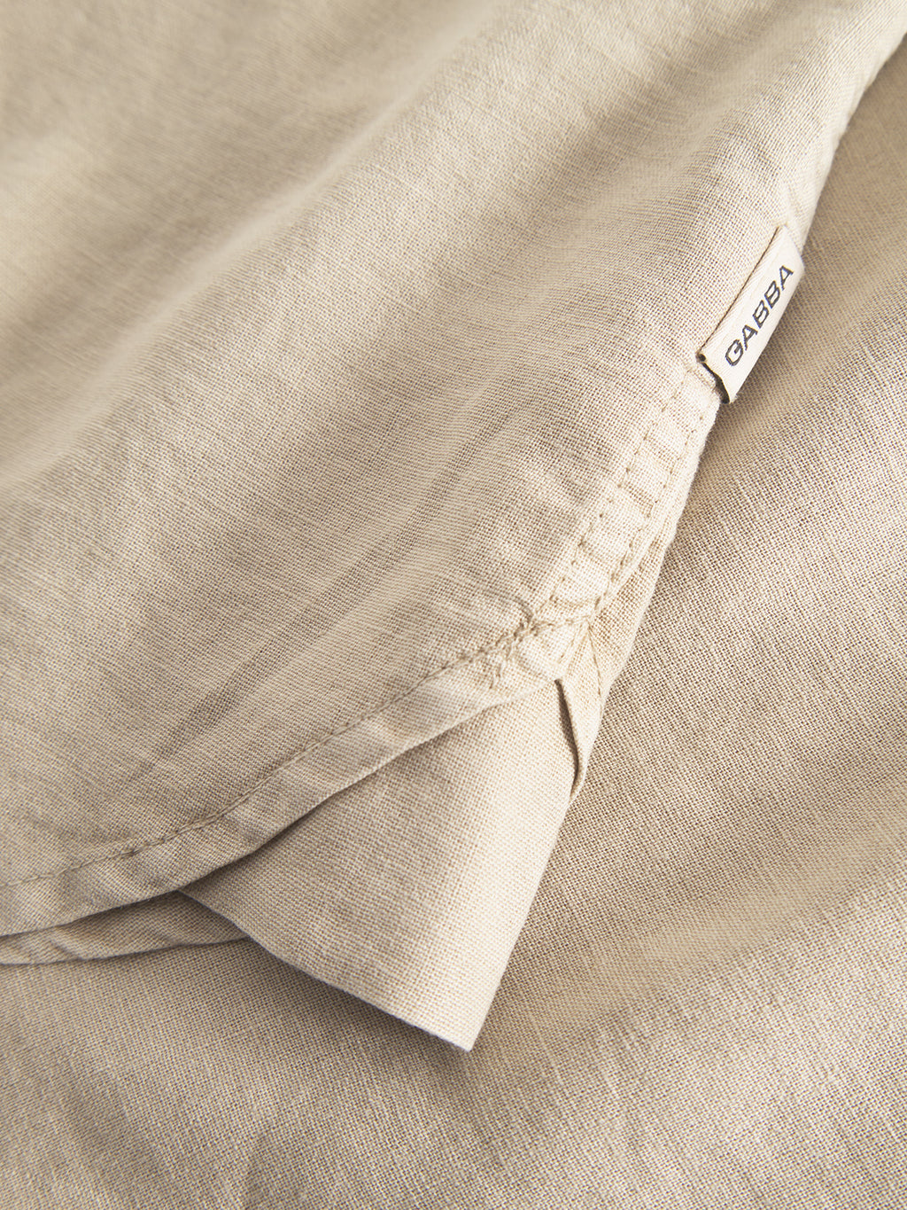 Close-up of layered beige linen fabric with visible stitching, evoking comfort. A small Gabba tag is near the edge, suggesting its from the Sven Reg Lino SS - Sand Plaza shirt. The fabrics smooth texture and gentle folds hint at its use in a premium design.