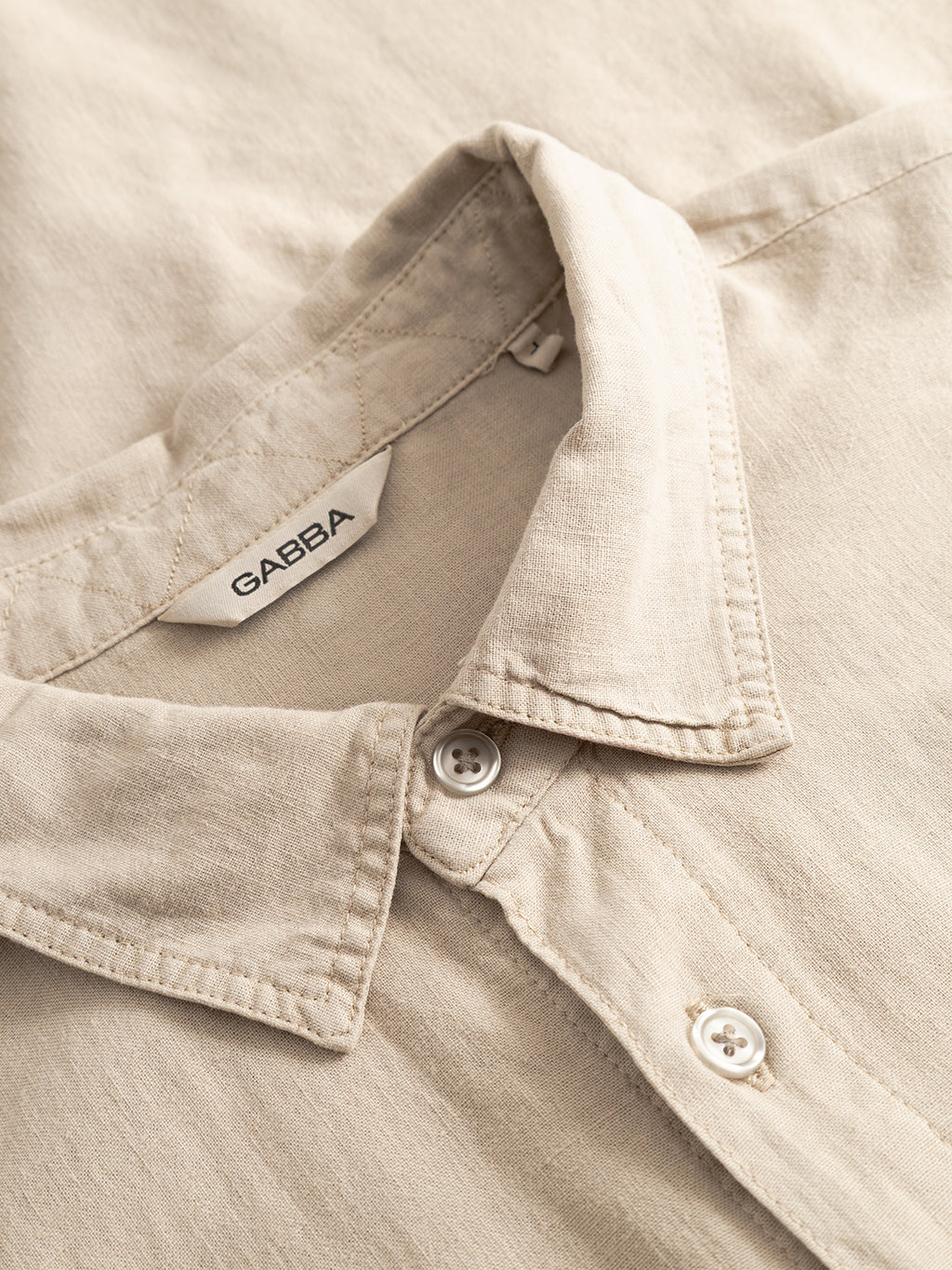 A close-up of the Sven Reg Lino LS - Sand Plaza shirt by Gabba shows its soft, durable beige fabric, white label with the Gabba logo, and neat fold with two white buttons.