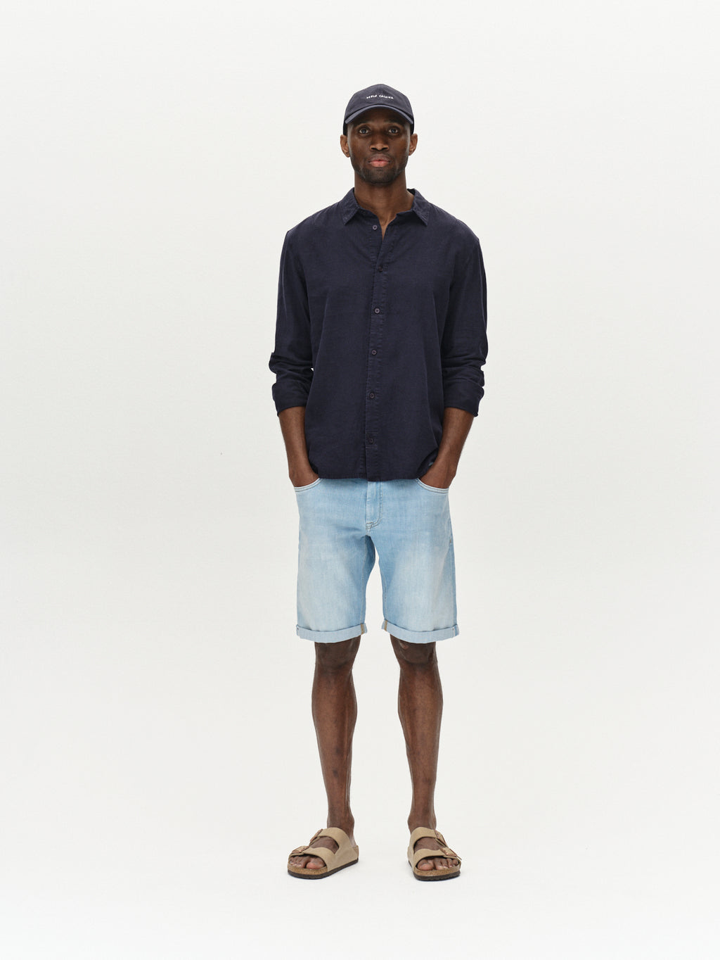 A man stands against a white background, hands in pockets, wearing the Gabba Sven Reg Lino LS - Night Sky shirt, light denim shorts, a baseball cap, and beige sandals.