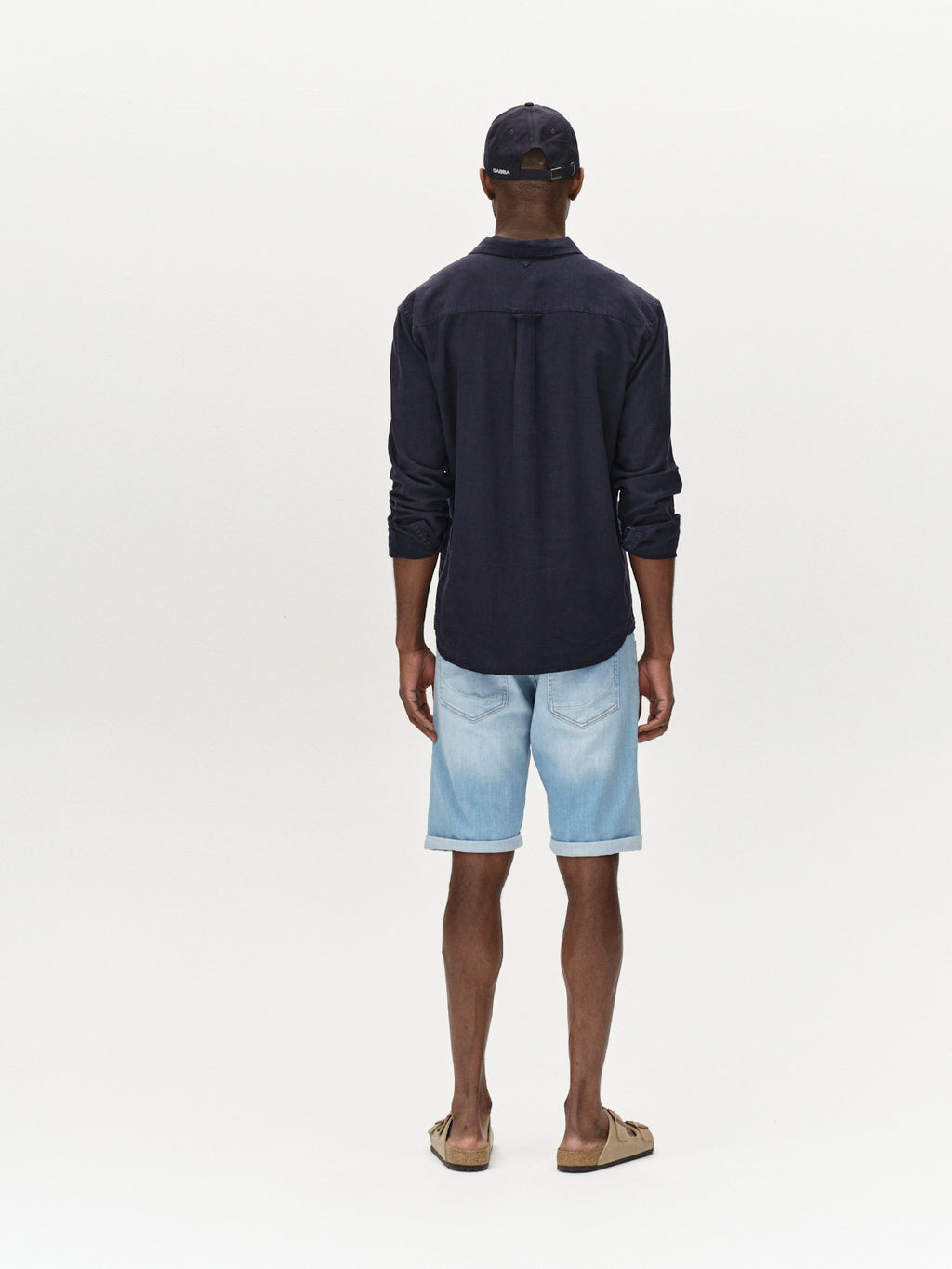 A man stands with his back to the camera, exuding comfort in a Gabba Sven Reg Lino LS - Night Sky shirt and light linen denim shorts with sandals. A dark cap rests snugly on his head against a plain, neutral background.