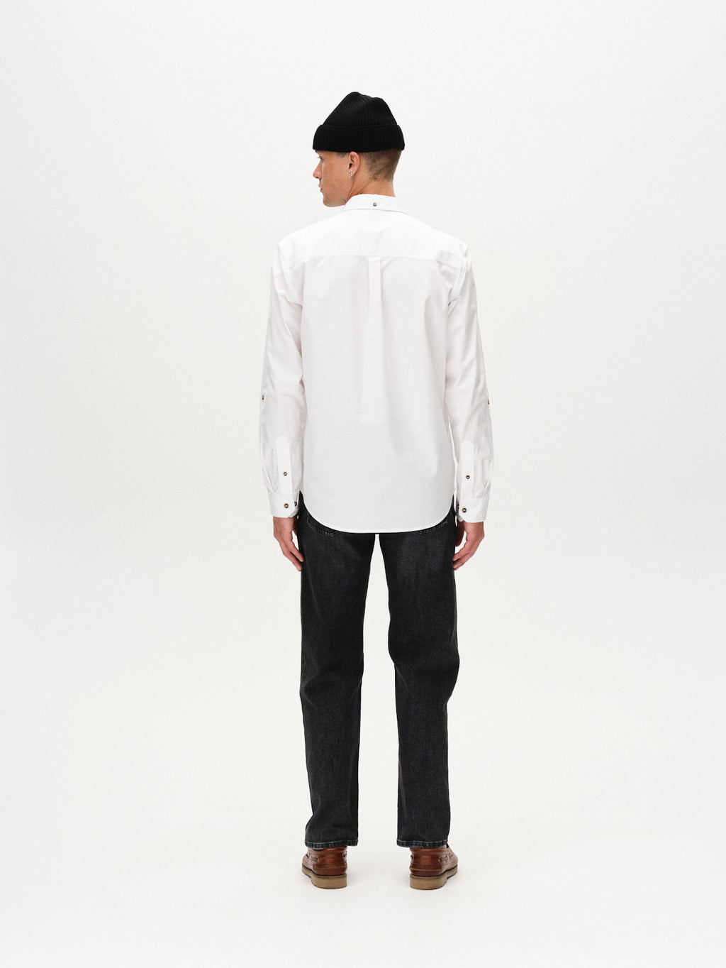 A person stands with their back to the camera in a white long-sleeve shirt, Gabba Max F1011 black denim jeans made with Wiser Wash technology, brown shoes, and a black beanie against a plain white background.
