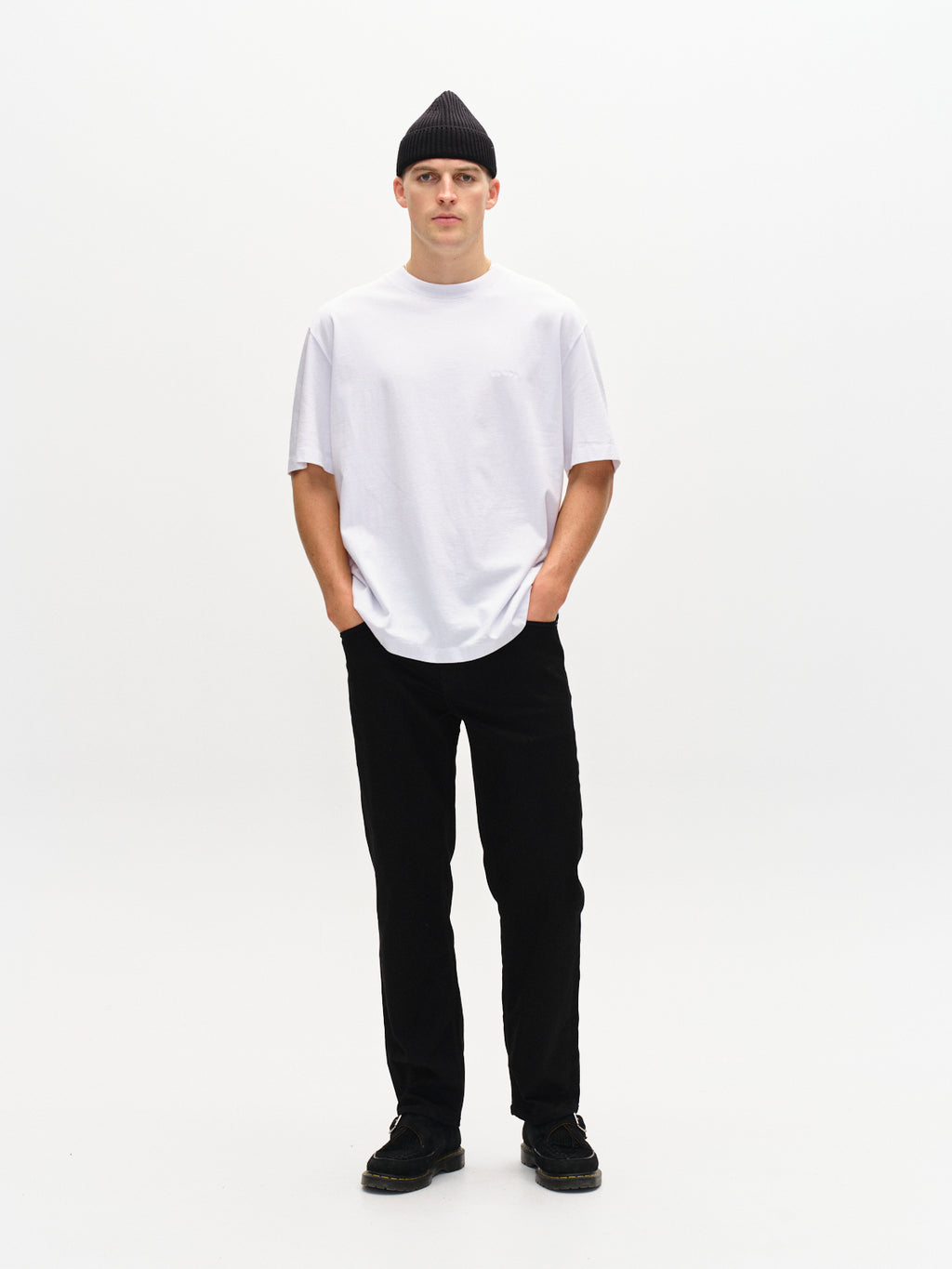 A person wearing a modern slim fit white t-shirt, Gabbas Max Sword black denim pants, black shoes, and a black beanie stands against a white background. Hands in pockets, they face the camera with casual confidence.
