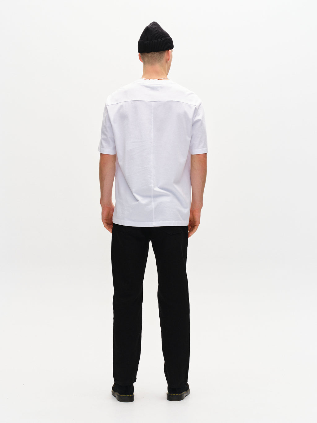 A person faces away from the camera, wearing a white T-shirt paired with Gabbas Max Sword - Black Denim jeans and sleek black wash shoes, topped off with a matching beanie. The minimalist setting features a plain white backdrop.