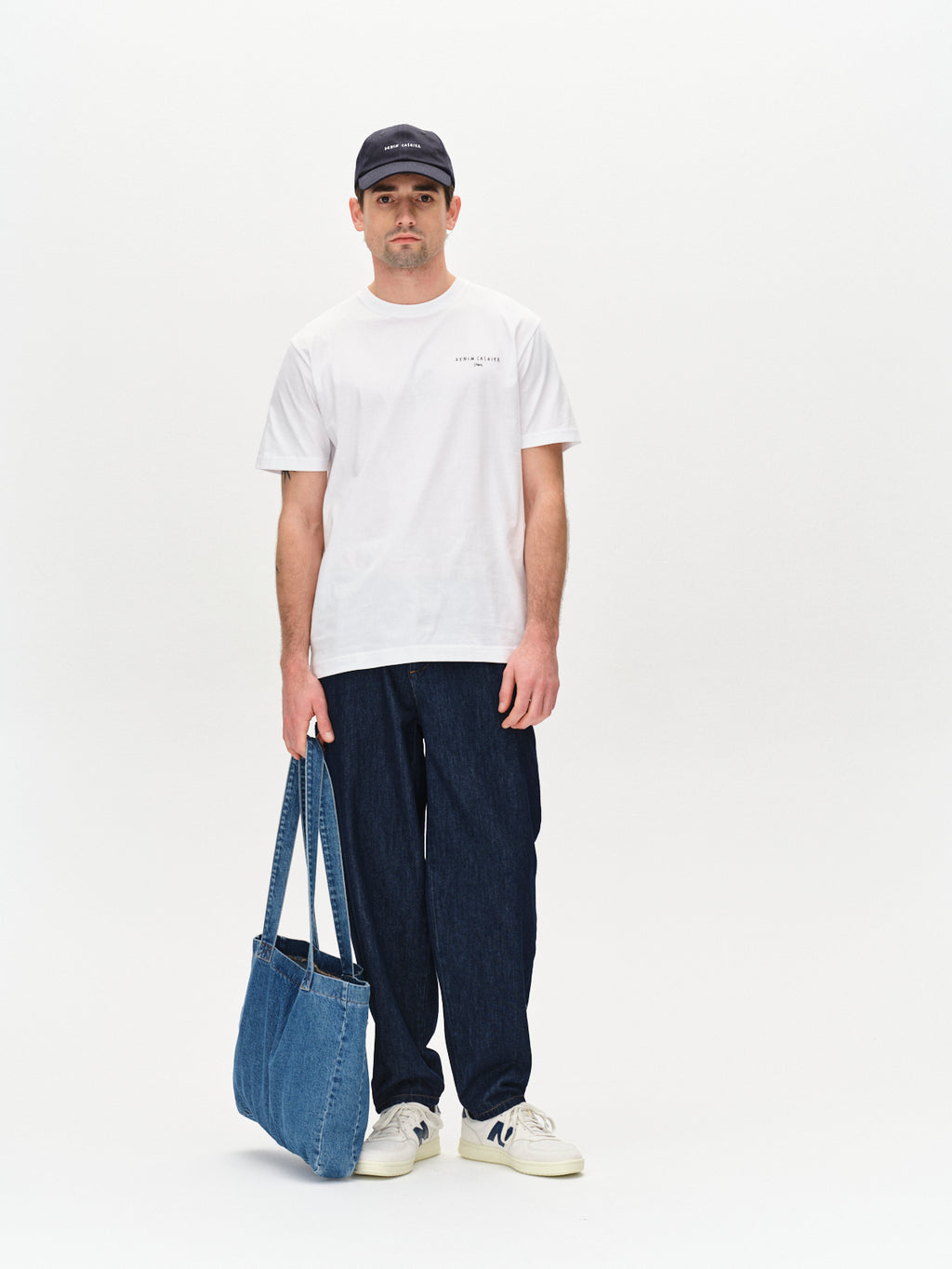 A man in a Gabba Dune Kiosk A SS GOTS white t-shirt, dark pants, and sleek white sneakers stands against a plain backdrop. He wears a bold seasonal print cap and holds a chic blue denim tote bag.