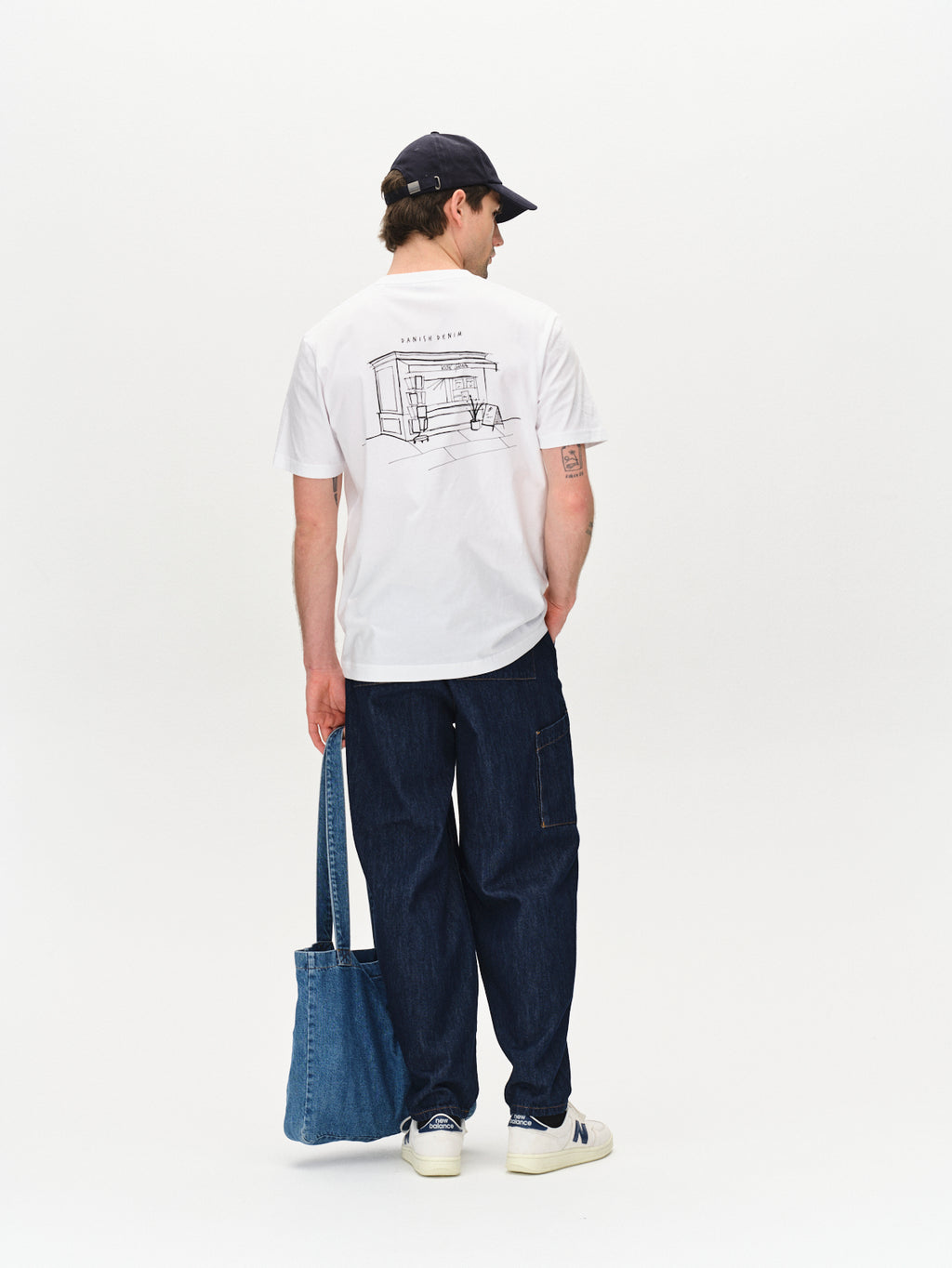 A man wears the Dune Kiosk A SS GOTS - White T-shirt by Gabba, featuring a line drawing on the back. He pairs it with dark jeans, white sneakers, and a black cap while holding a denim tote bag, standing slightly turned against a plain white background.