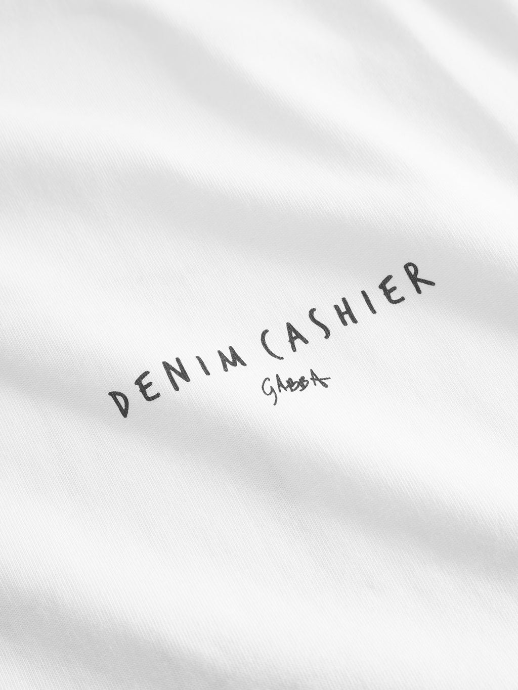 Close-up of a white fabric labeled Dune Kiosk A SS GOTS - White by Gabba, featuring DENIM CASHIER. Crafted from organic cotton with a signature-like mark in black, showcasing the striking texture of the background fabric.