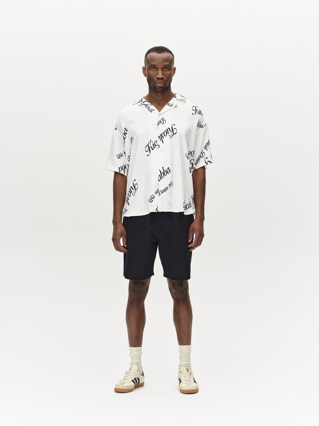 A man wears the Gabba Jens Over Resort Kiosk SS - Navy Pattern shirt, styled with black shorts, beige socks, and beige sneakers with black stripes, standing before a plain white background.