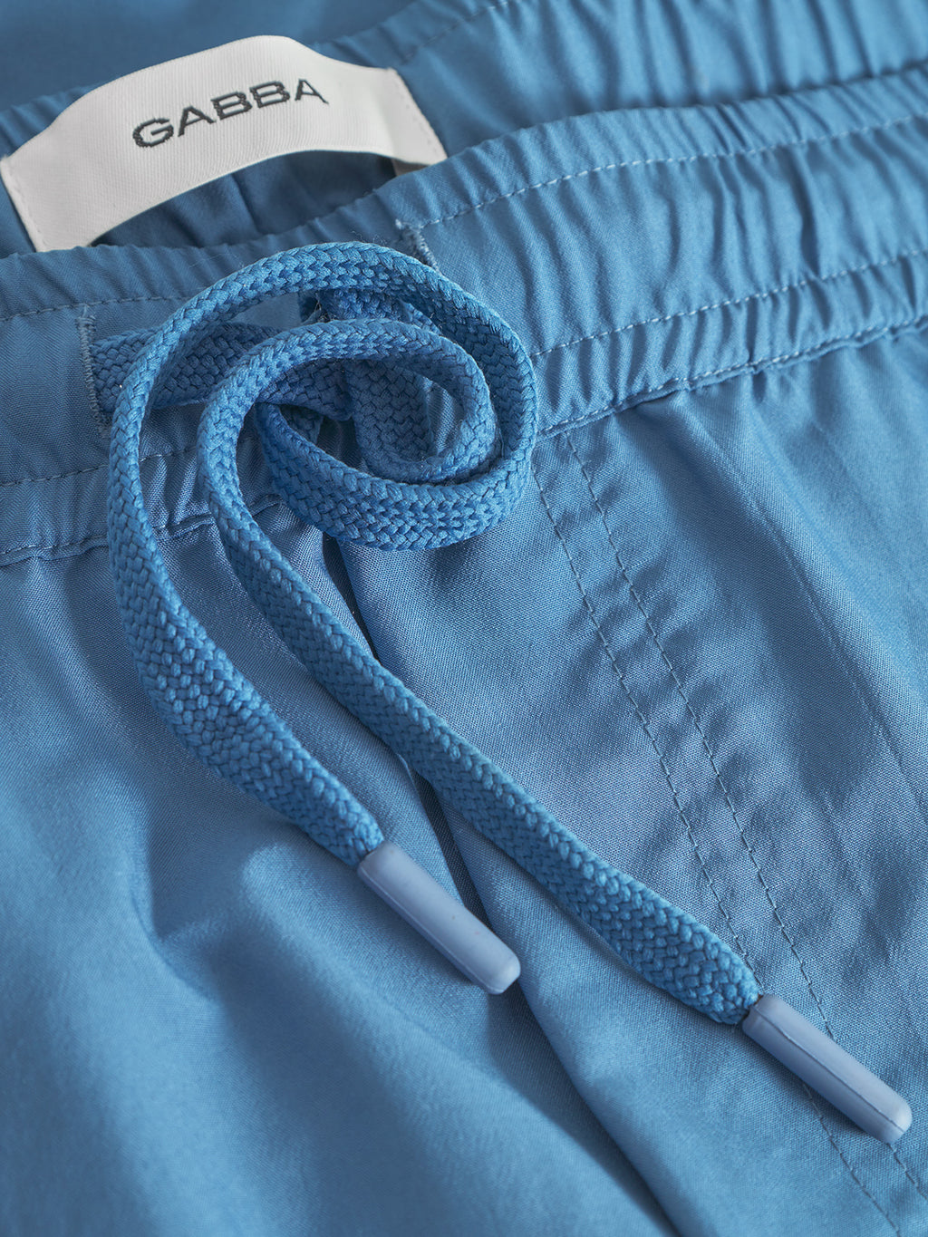 Close-up of Jackson Plain - Blue Horizon swim shorts by Gabba, showcasing a braided blue drawstring, elastic waistband, and side pockets. A white Gabba label is partly visible inside the waistband.