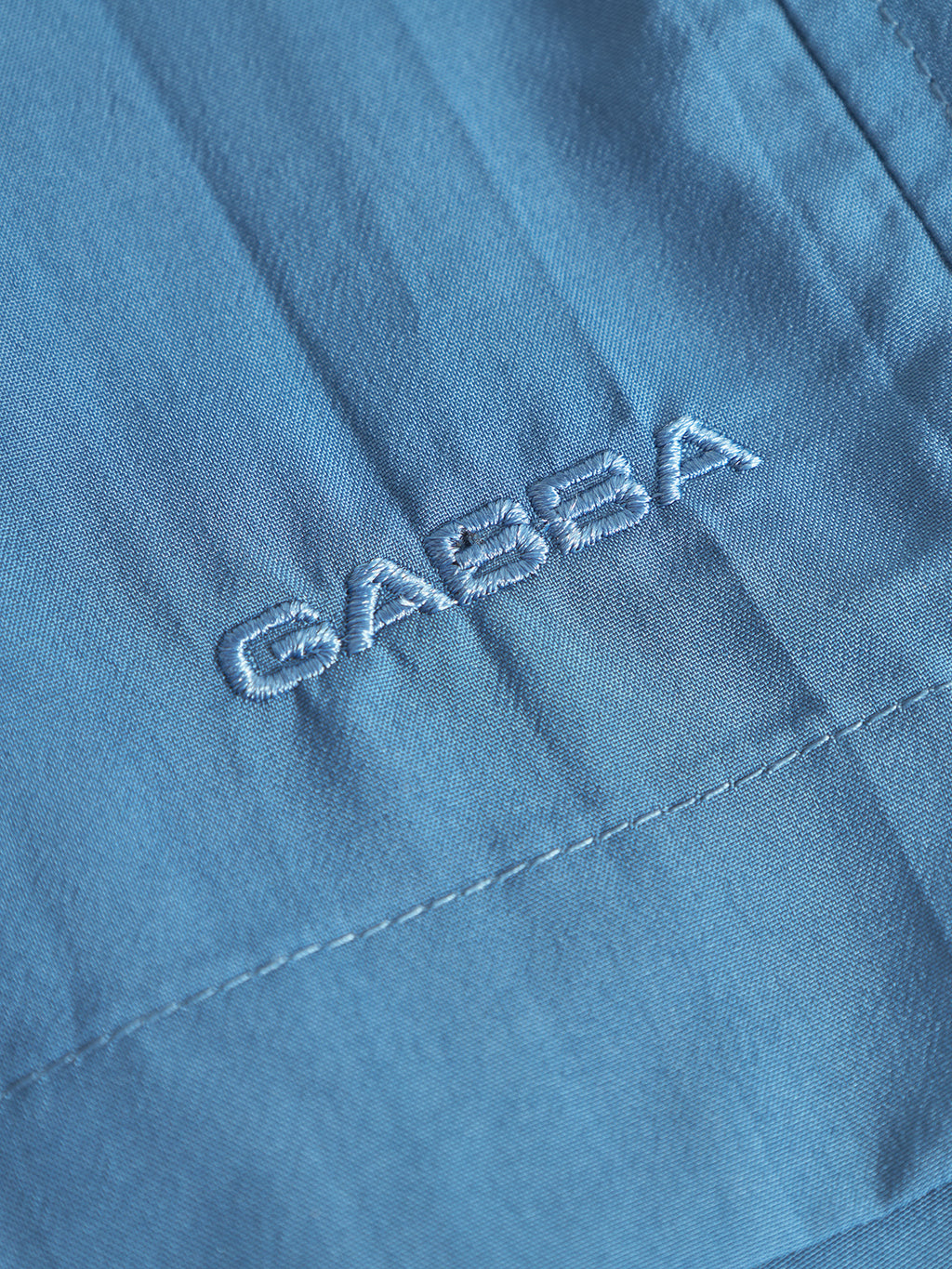 Close-up of Gabba Jackson Plain - Blue Horizon swim shorts fabric with Gabba embroidered in matching blue thread. The stitching, seams, and elastic waistband details are clearly visible.