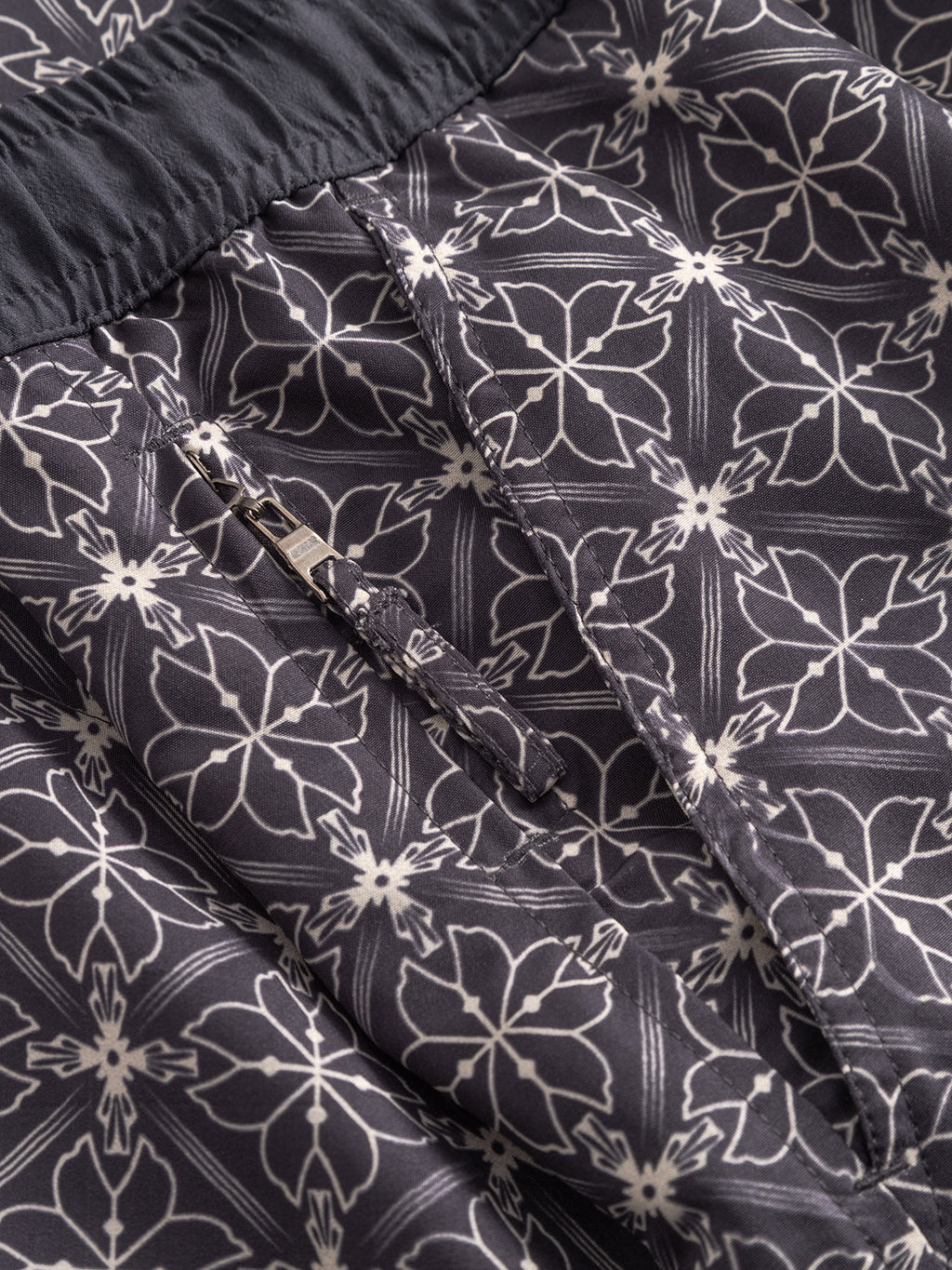 Close-up of Gabbas Jackson Print - Multi Pattern swim shorts, showcasing a white floral geometric design on dark fabric, with an elastic waistband, side pockets, and a small zippered pocket.