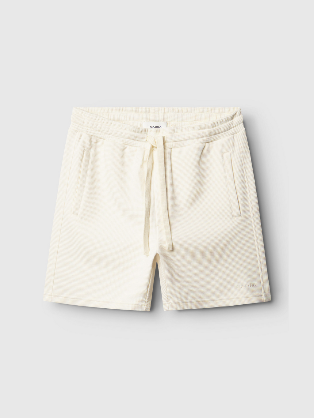 Gabba Tim Waff - Bone White shorts in off-white waffle fabric with elastic waistband, adjustable drawstring, and side pockets. Subtle branding near the hem. Shown on a plain light gray background for premium comfort and style.