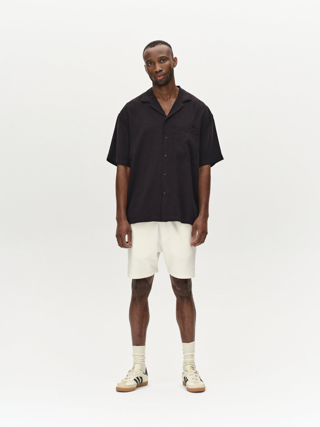 A man stands against a plain background, dressed in a black short-sleeve button-up shirt and premium Tim Waff - Bone White shorts by Gabba, made from waffle fabric. He completes his relaxed look with beige sneakers featuring black stripes, showcasing comfort and durability while gazing slightly forward.
