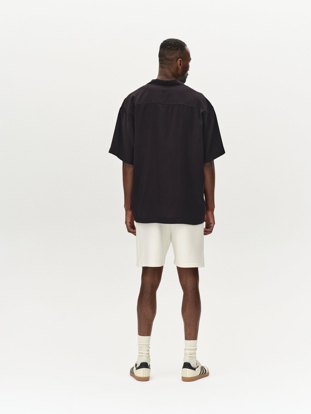 A person stands facing away, highlighting comfort and durability in a black short-sleeve shirt, premium Tim Waff - Bone White shorts by Gabba, white socks, and brown shoes with black stripes against a plain white background.