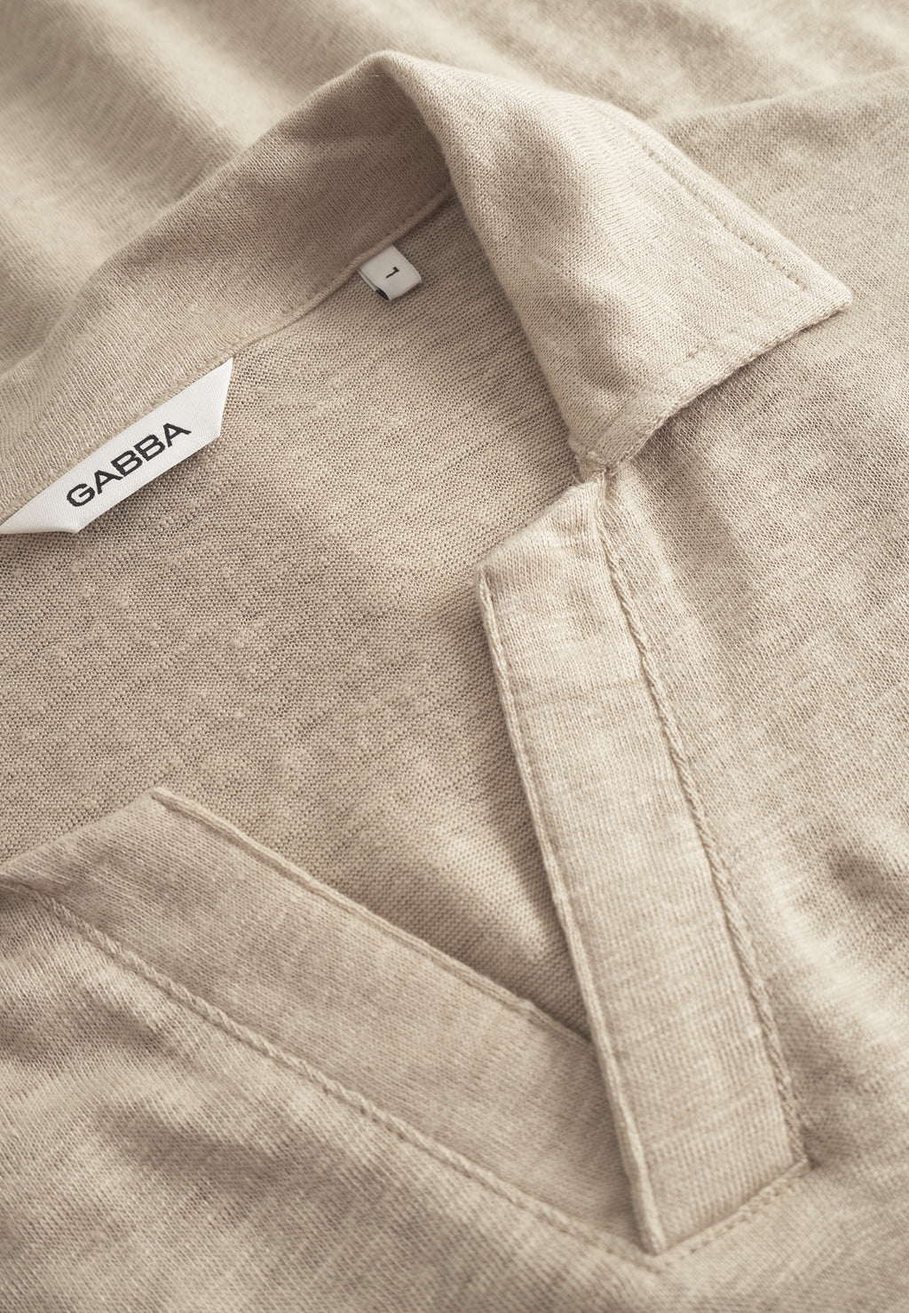 Close-up of the Gabba Trevor - Sand Plaza shirt, showcasing a beige collared design with a deep V-neck for superior comfort. A small white tag displays Gabba. The shirt is folded to highlight its fabric texture, exuding refined elegance.