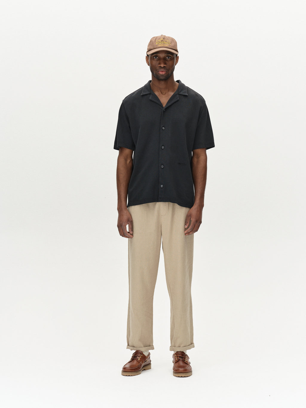 Against a plain white backdrop, a person exudes ultimate comfort in Gabbas Neo Pique Resort SS in Meteorite, paired with beige pants, brown shoes, and a beige cap. Their neutral expression and relaxed arms at their sides enhance the serene vibe.