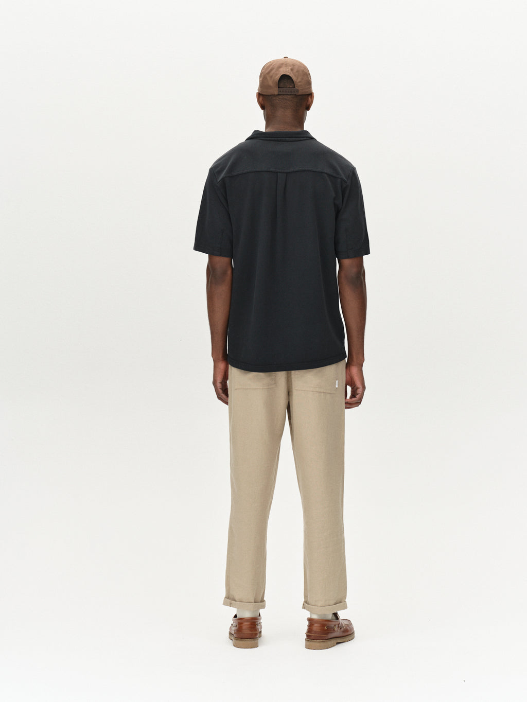 Against a plain white backdrop, someone embraces their style in ultimate comfort wearing Gabbas Neo Pique Resort SS in Meteorite, complemented by beige pants, brown shoes, and a tan cap, with hands relaxed at their sides.