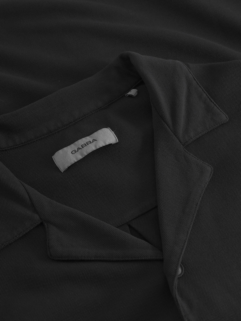 A close-up of the Neo Pique Resort SS - Meteorite by Gabba reveals a black collared shirt with a white label reading GABBA. The textured fabric and slightly turned-up collar offer ultimate comfort and casual elegance, showcasing its versatile appeal for personal style.