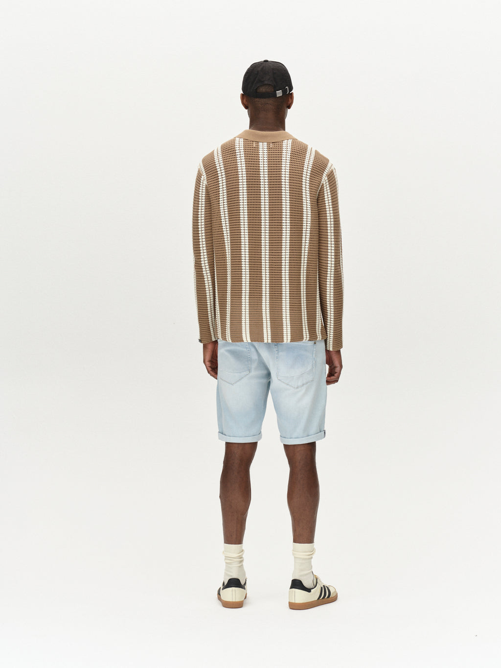 A person facing away from the camera wears Gabbas Jason K3787 SANZA Shorts in light blue denim, a black cap, a brown and white striped sweater, white socks, and brown sneakers with black stripes against a plain white background.