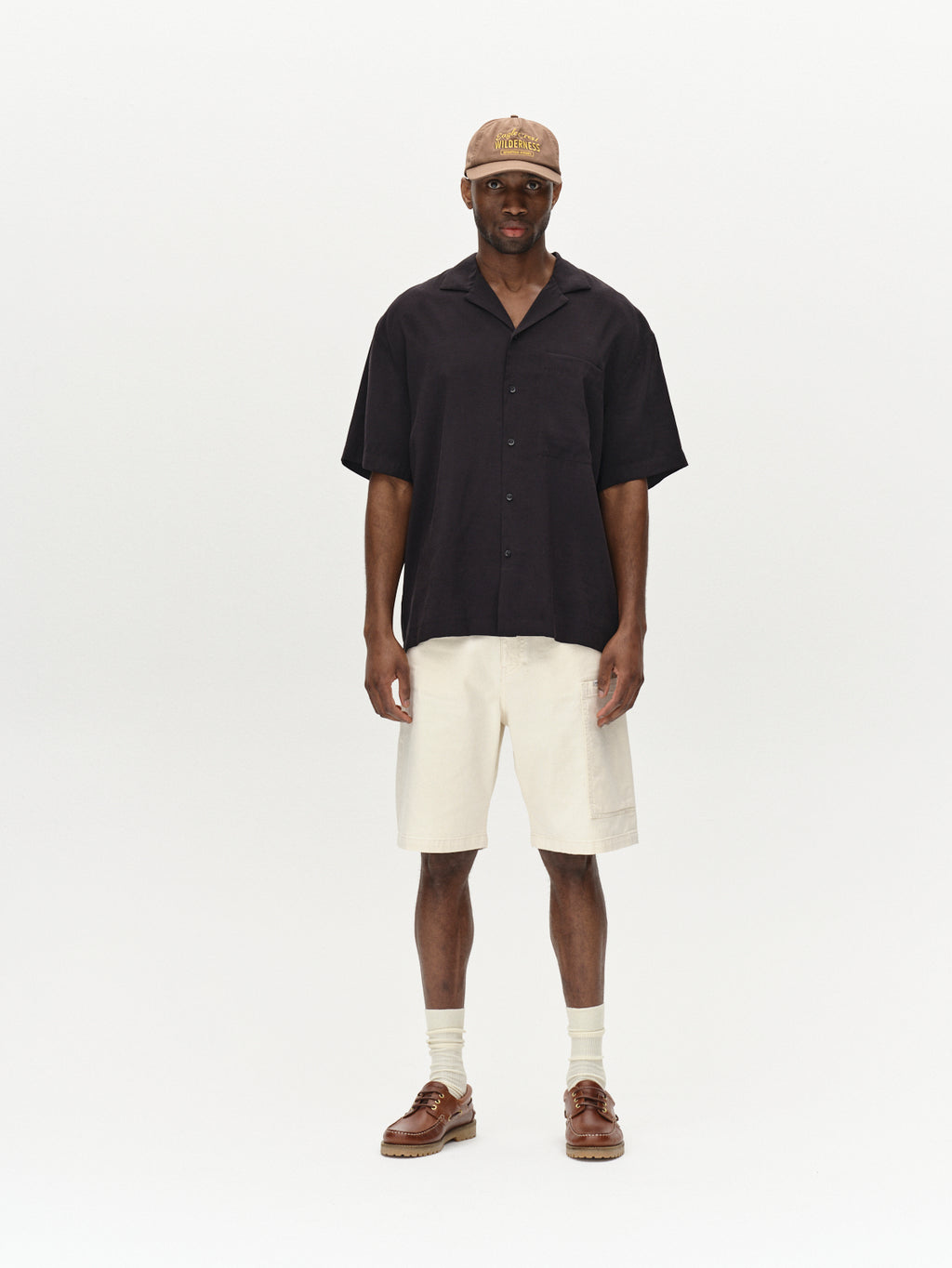 A person stands against a plain background wearing a brown cap, short-sleeved black button-up shirt, Gabbas Maxwell Luis Cargo Shorts - Ecru, and brown leather shoes with beige socks.