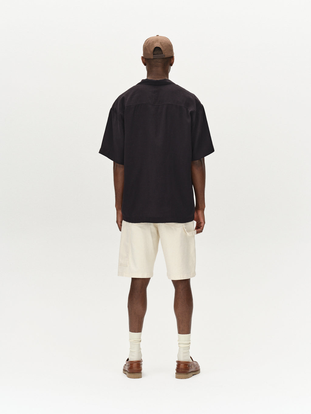 A person faces away, wearing a black oversized T-shirt, Gabbas Maxwell Luis Cargo Shorts in ecru, white socks, brown sandals, and a beige cap. The backdrop is a neutral light color with an understated workwear vibe.