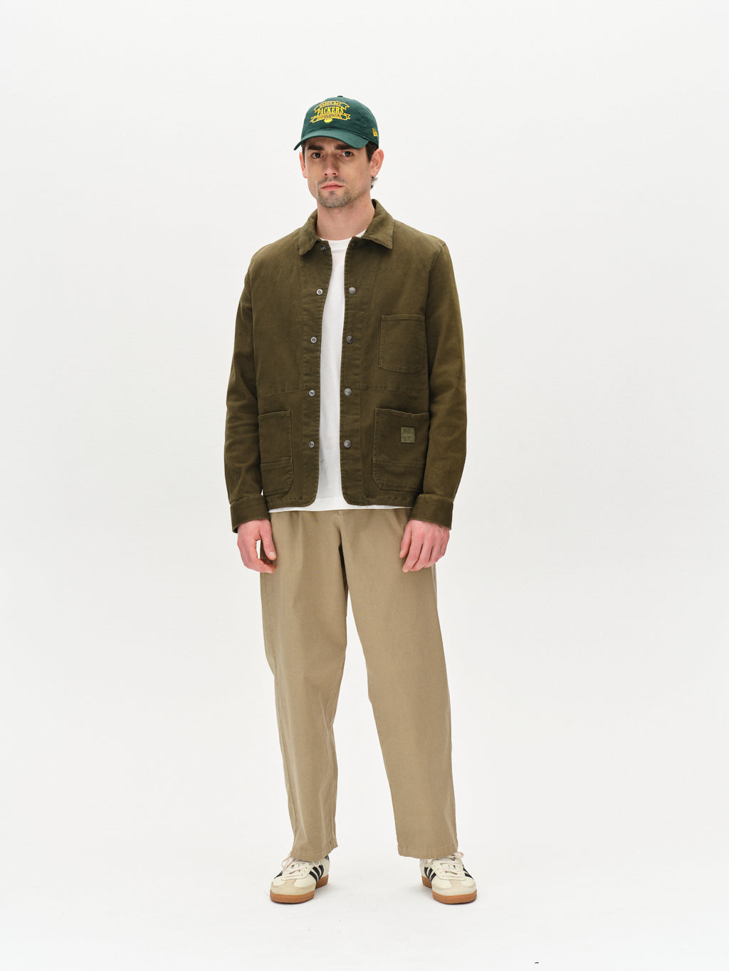 A man stands against a plain white background, wearing Gabbas Beta Twill - Army overshirt with a green cap, white shirt, beige pants, and white sneakers.