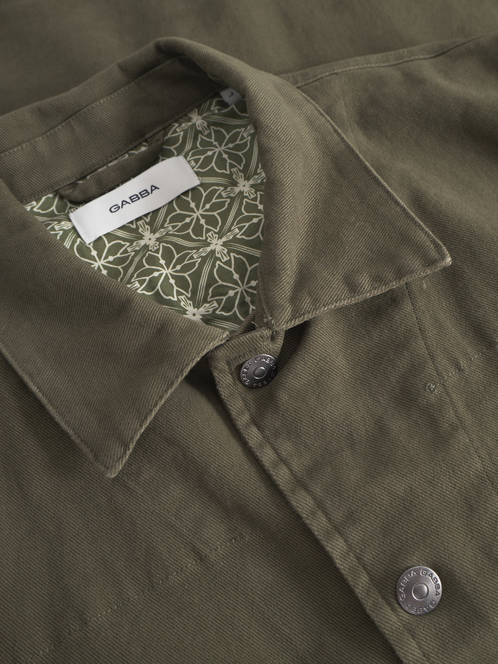 Close-up of the Gabba Beta Twill - Army jacket, an olive green workwear-inspired overshirt with silver buttons, a patterned green and white inner lining, and a Gabba brand label sewn inside the collar.