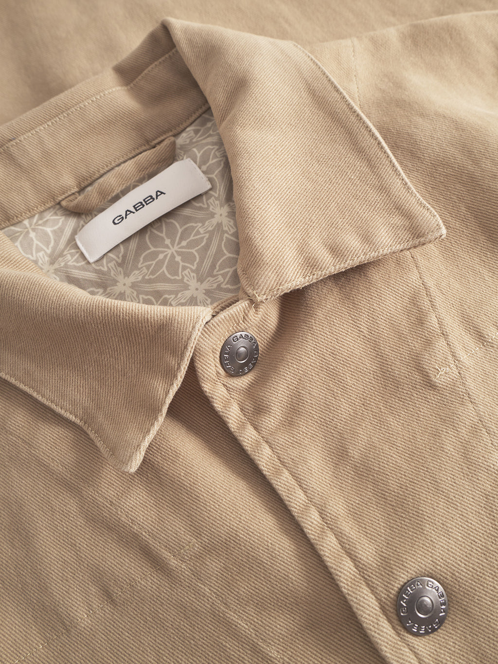 A close-up of the Gabba Beta Twill - Sand overshirt with metal buttons and a white Gabba label. The workwear-inspired design showcases a patterned inner lining visible near the collar.