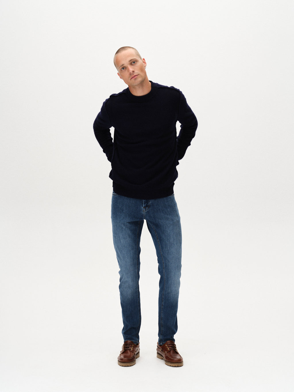 A man stands against a plain white background, wearing a dark long-sleeve sweater, Gabbas Jones Vanew - Mid Blue Denim jeans with a slim super-stretch fit, and brown shoes, his hands behind his back and a neutral expression.